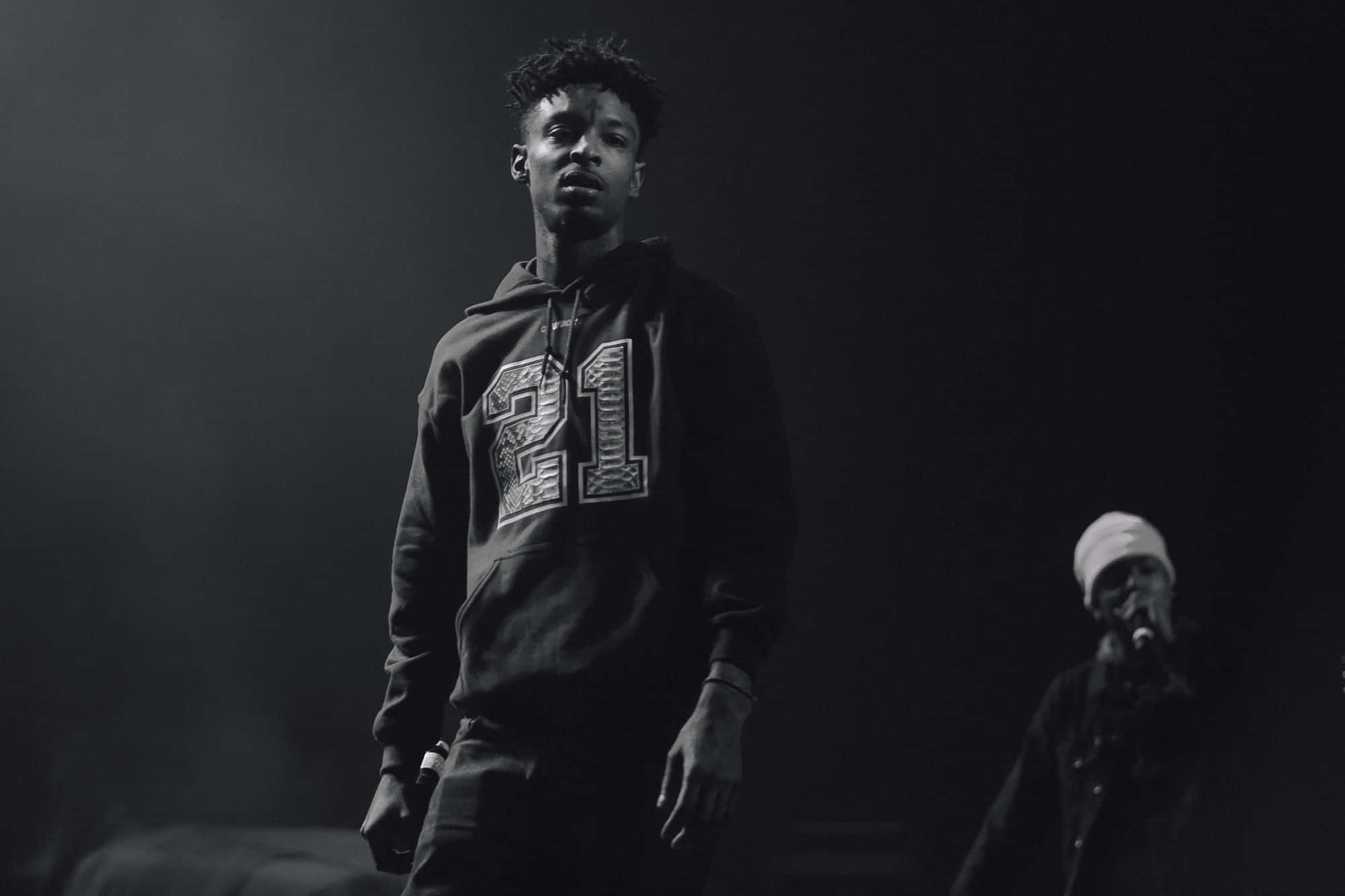 亞特蘭大饒舌歌手 21 Savage 登台表演 壁紙 — 一張黑白照片，一名身穿印有「21」連帽衫的年輕男子在表演期間站在燈光昏暗的舞台上。