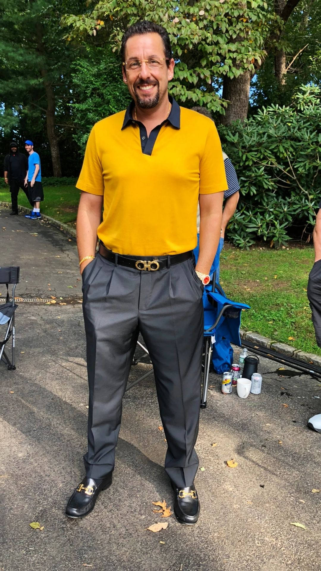 亞當·桑德勒正式擺姿 壁紙 — 一位微笑的男子身穿黃色Polo衫和灰色長褲，自信地站在戶外鋪砌的小徑上，周圍環繞著茂密的綠樹和樹葉。