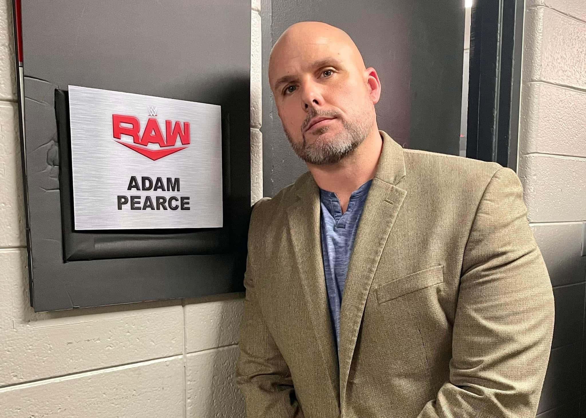 亞當·皮爾斯 WWE 節目嘉賓 壁紙 — 光頭山羊鬍鬚男穿著米色西裝外套站在RAW摔角辦公室門牌旁邊。