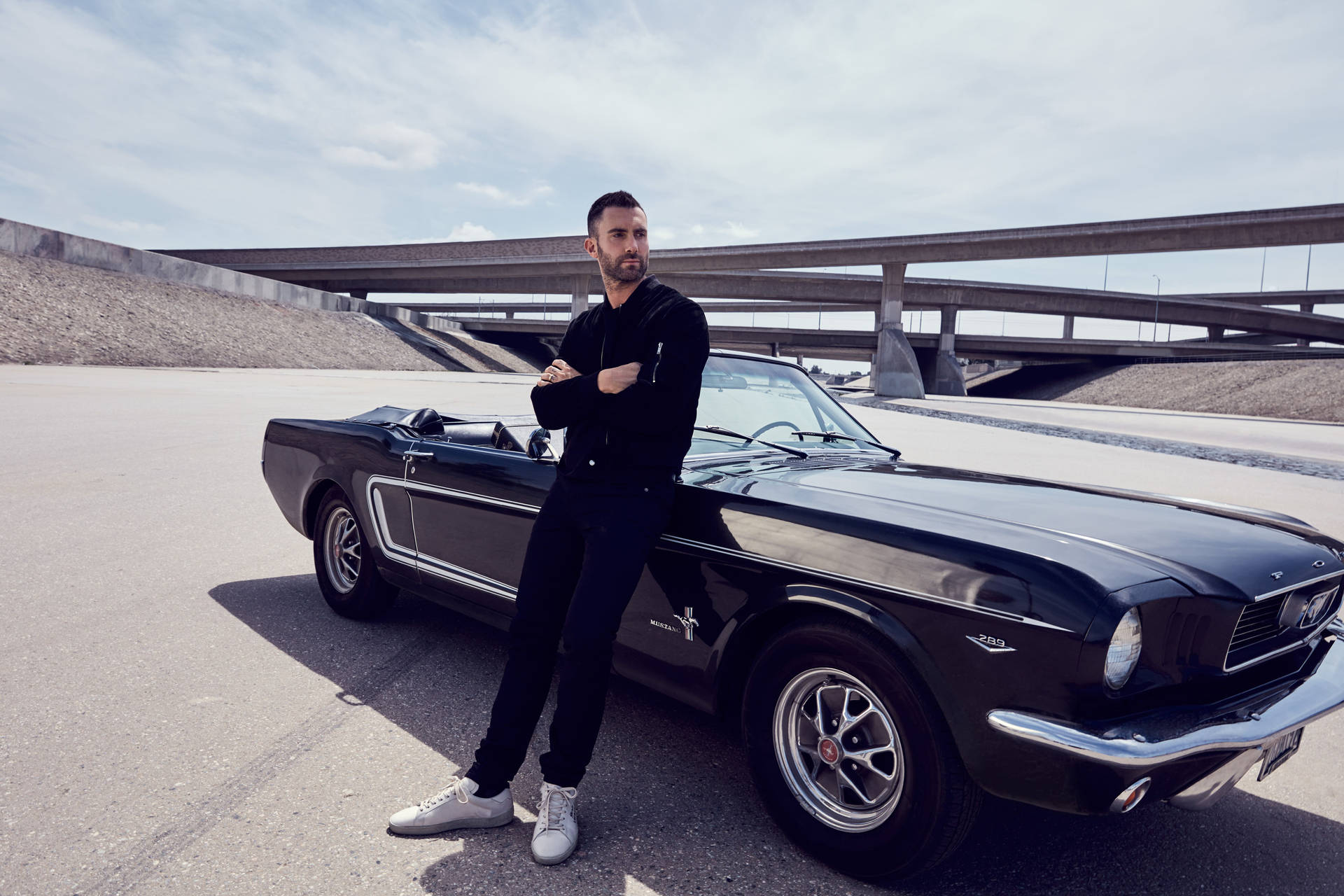 男子靠在一輛黑色經典Mustang開篷車旁，街道上有高架橋。