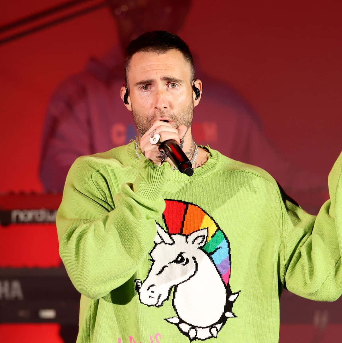 亞當·萊文在舞台上搖滾演出 壁紙 — Adam Levine 在舞台上表演，身穿一件帶有彩虹獨角獸圖案的亮綠色毛衣。
