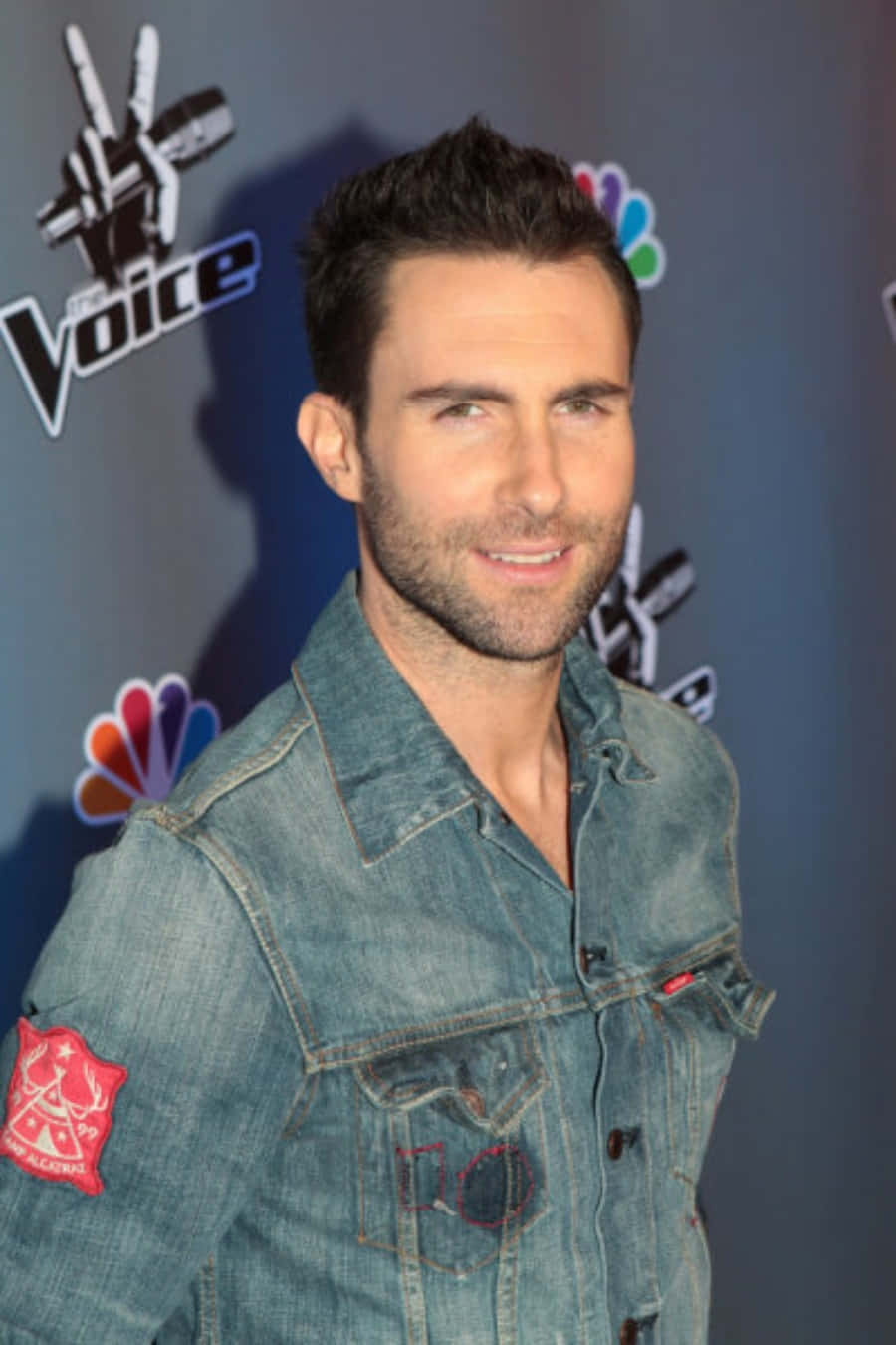 亞當·萊文現場演唱會 壁紙 — Adam Levine 身穿牛仔外套，在背後有NBC標誌的The Voice活動中自信微笑。