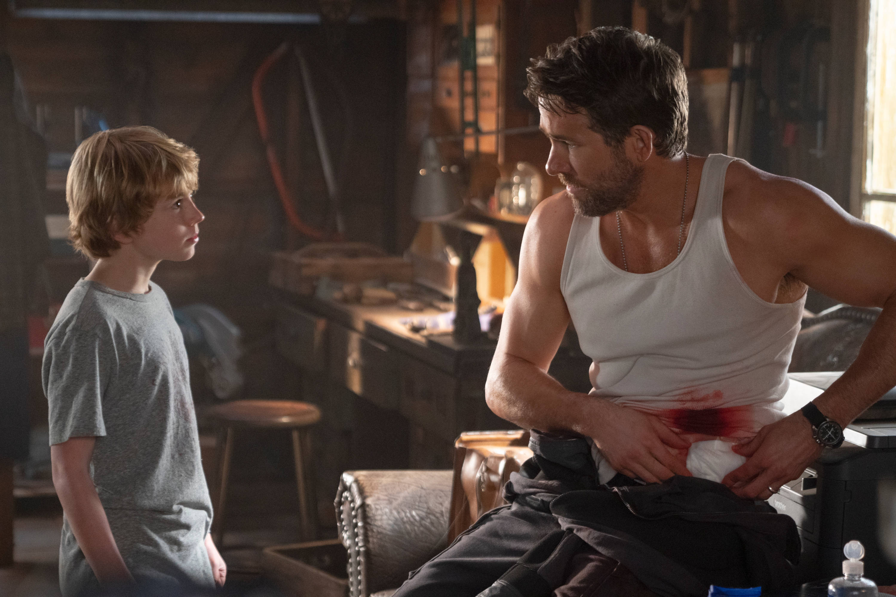 《亞當計劃》血腥畫面 壁紙 — 一個臀部有血淋淋傷口的男人坐在雜亂的工作室裏，一個小男孩專注地看着他。