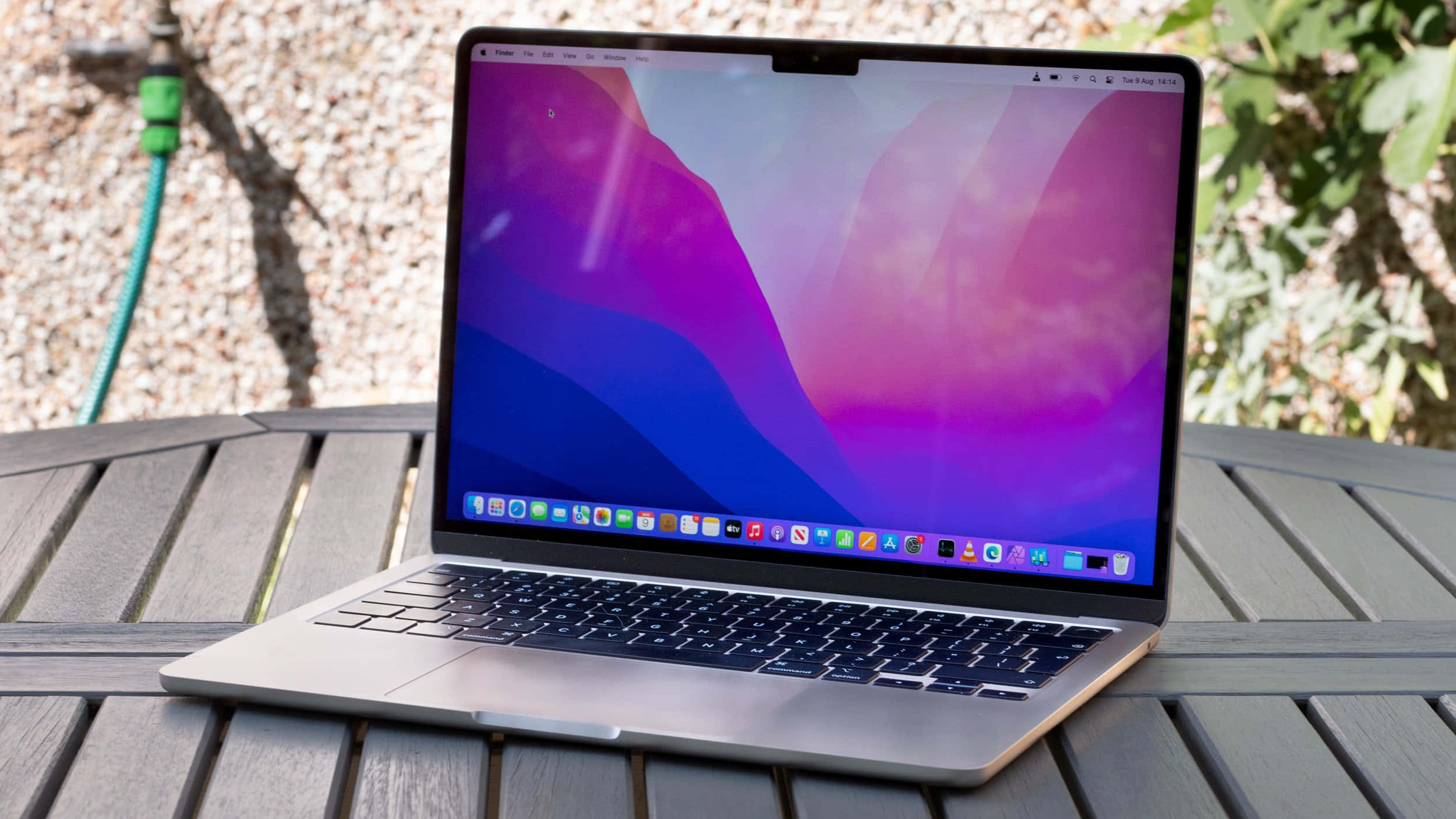 享受 MacBook 的絢麗 Retina 顯示屏 壁紙 — 一部銀色手提電腦，帶有充滿活力的紫色和藍色抽象壁紙，打開放在戶外的木條桌上。