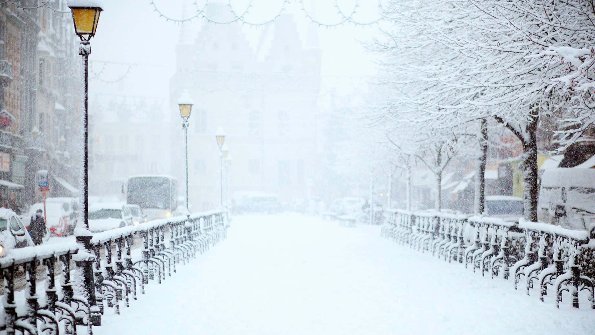 享受「雪景放大」的冬季美景 壁紙 — 大雪紛飛下，一條被雪覆蓋的城市街道，兩旁有復古路燈和積滿雪的樹木。
