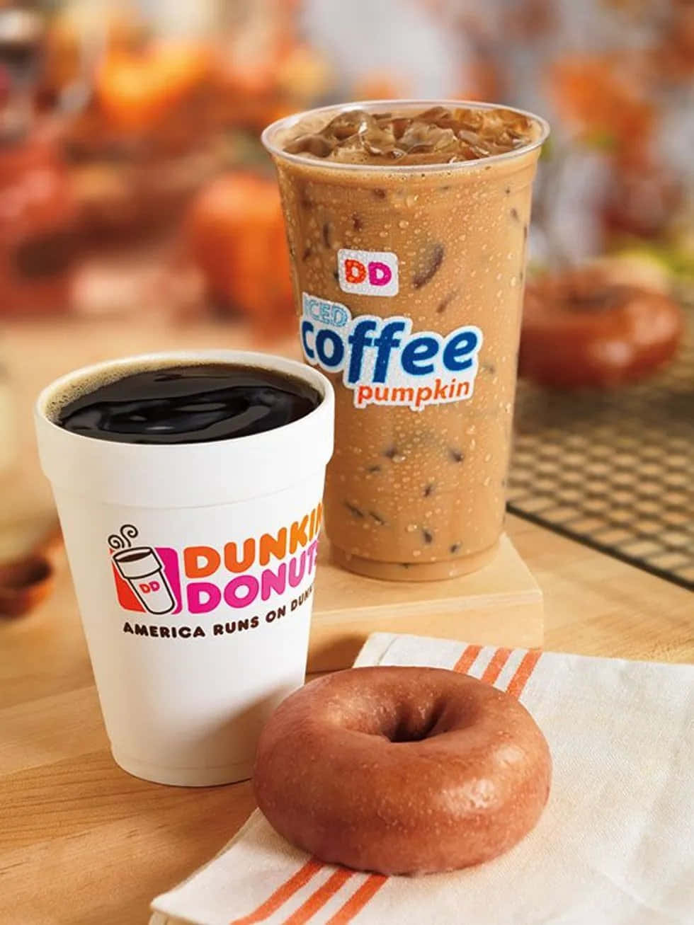 享受Dunkin' Donuts新鮮沖煮咖啡的美味！ 壁紙 — 一杯Dunkin' Donuts南瓜冰咖啡和黑咖啡放在木桌上，旁邊有一個糖霜冬甩，背景有秋葉。