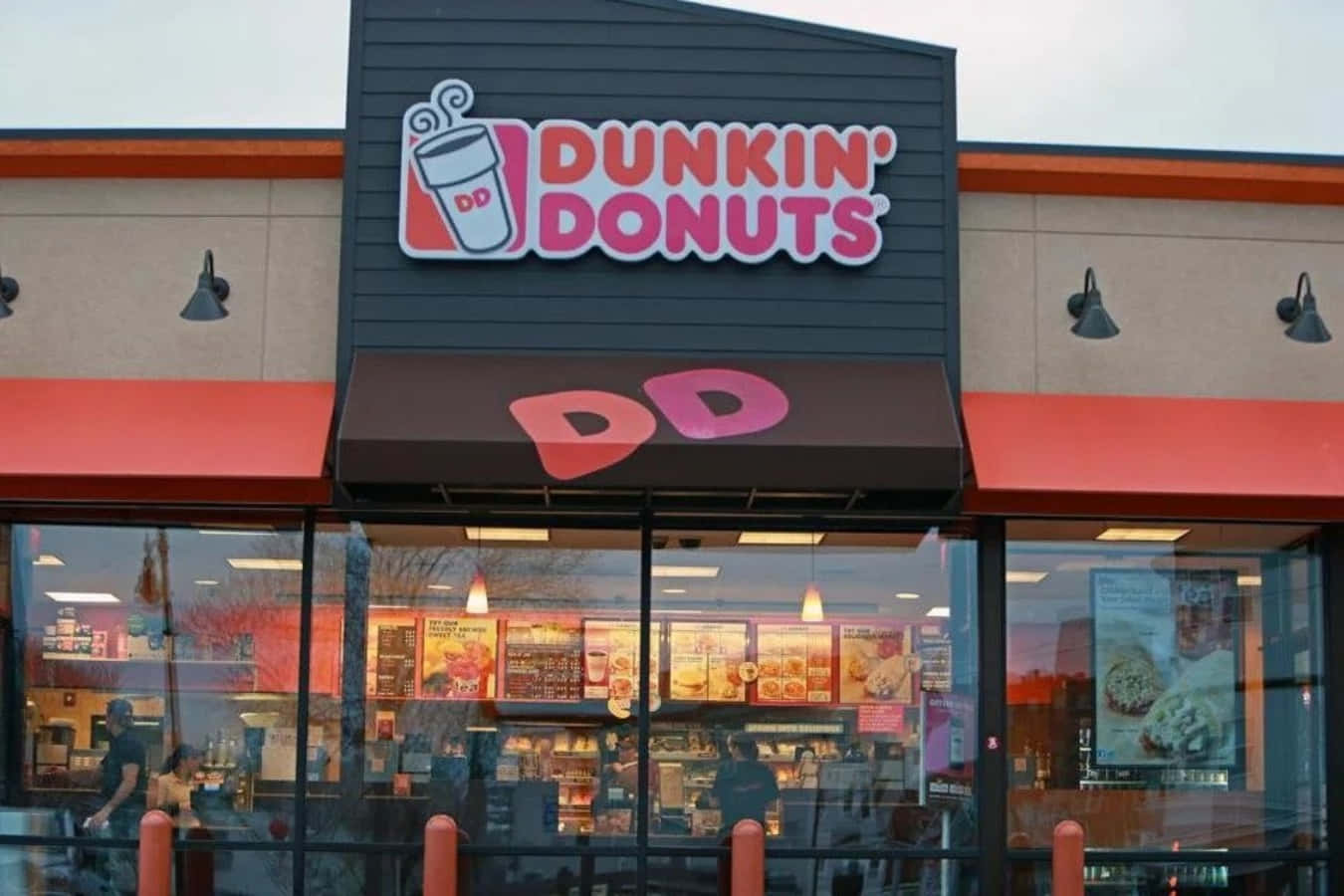 享受Dunkin' Donuts的美味 壁紙 — Dunkin' Donuts 店舖外觀，採用大膽的紅色和黑色品牌標誌，大型玻璃窗反射著多雲的天空和室內燈光。