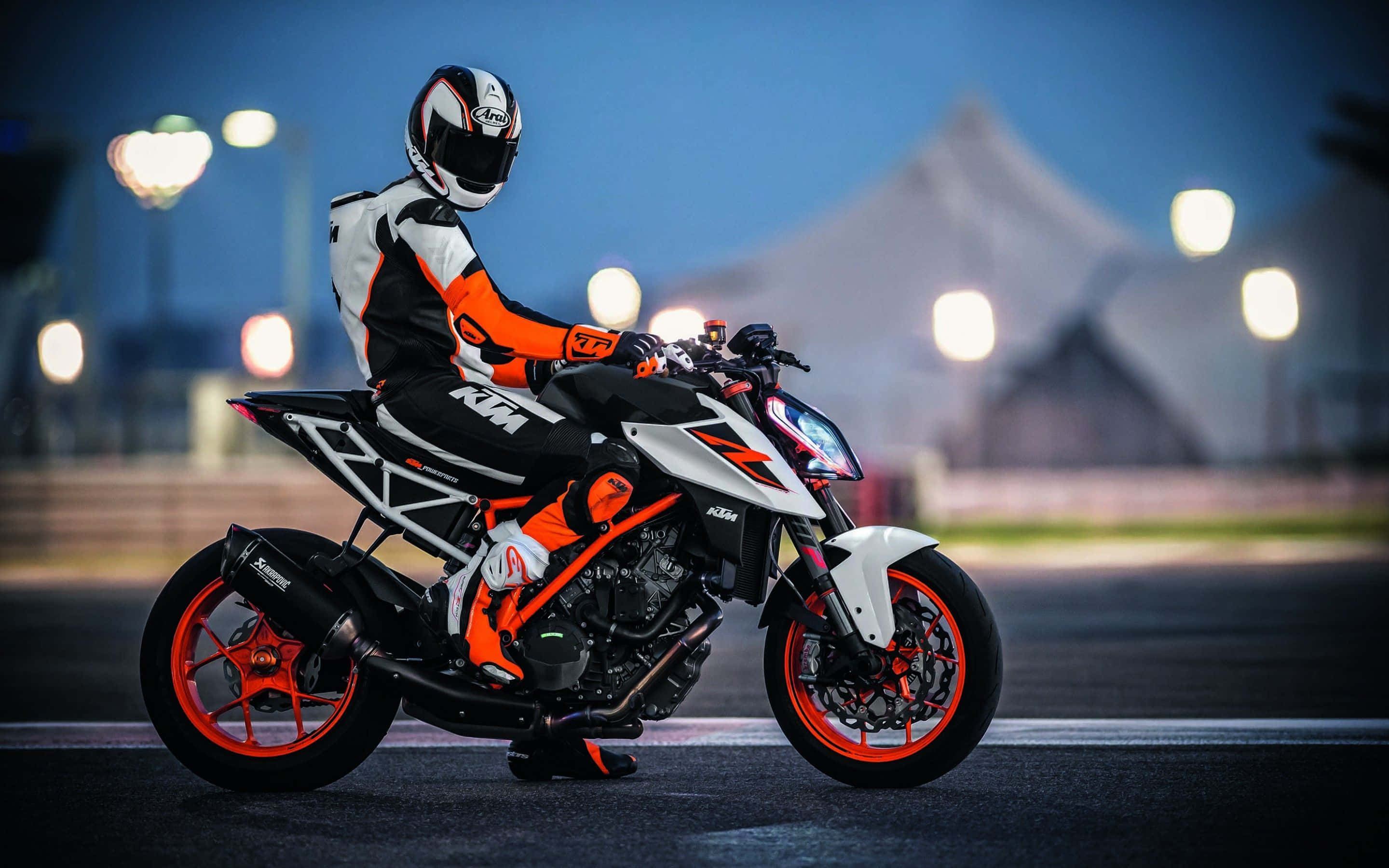 享受KTM Duke機車的刺激樂趣 壁紙 — 一名全副裝備的騎士在暮色城市燈光下，坐在一輛醒目的橙色和白色KTM電單車上。