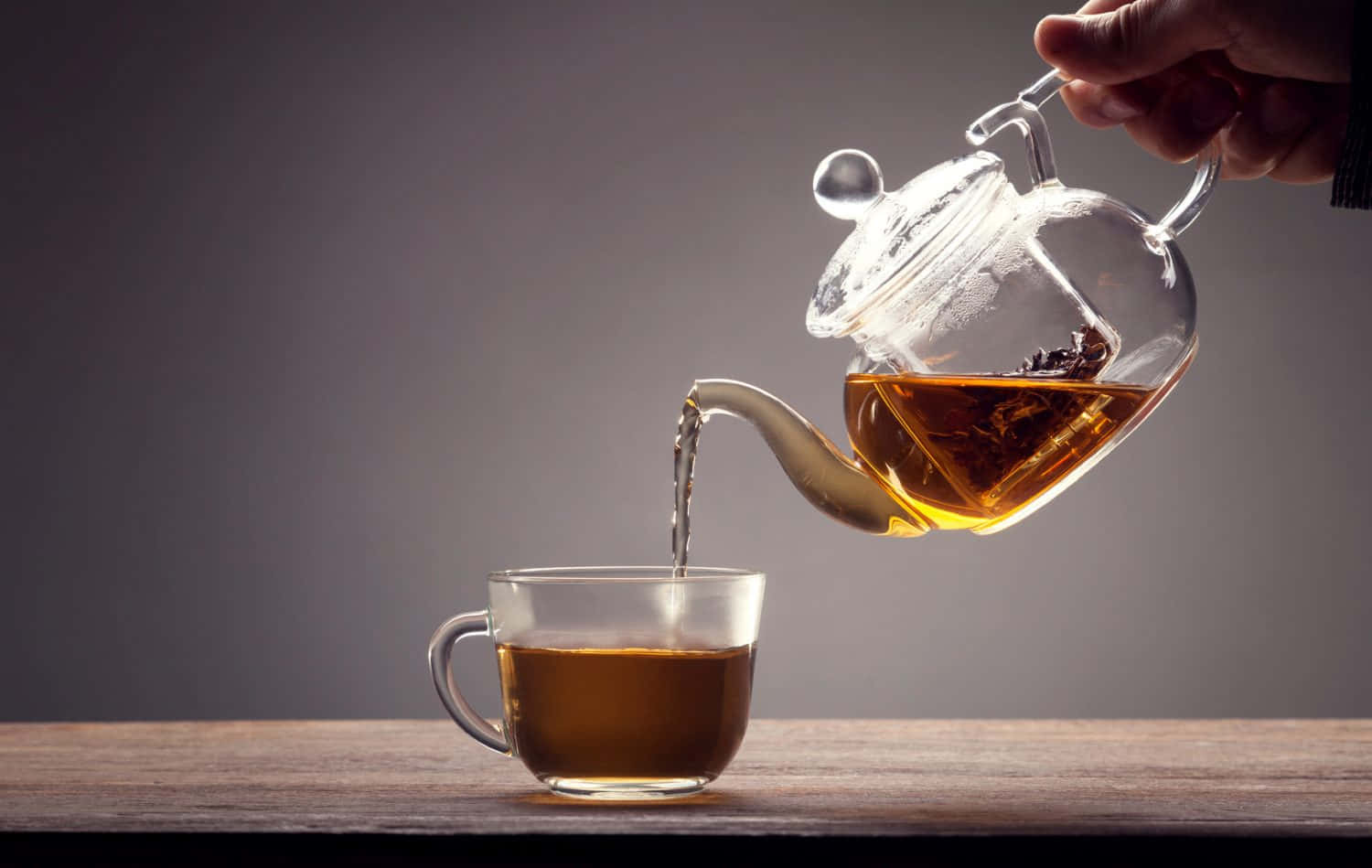 手將琥珀色茶從玻璃茶壺倒入透明杯中，木桌。