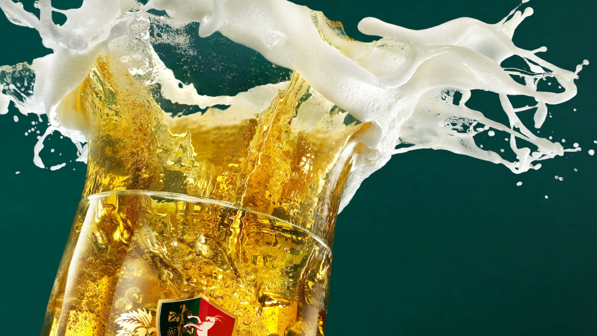 享受冰涼啤酒的輕盈美味 壁紙 — 金黃色的啤酒從玻璃杯中噴湧而出，帶有泡沫豐富的白色泡沫，在深藍綠色背景上戲劇性地飛濺。