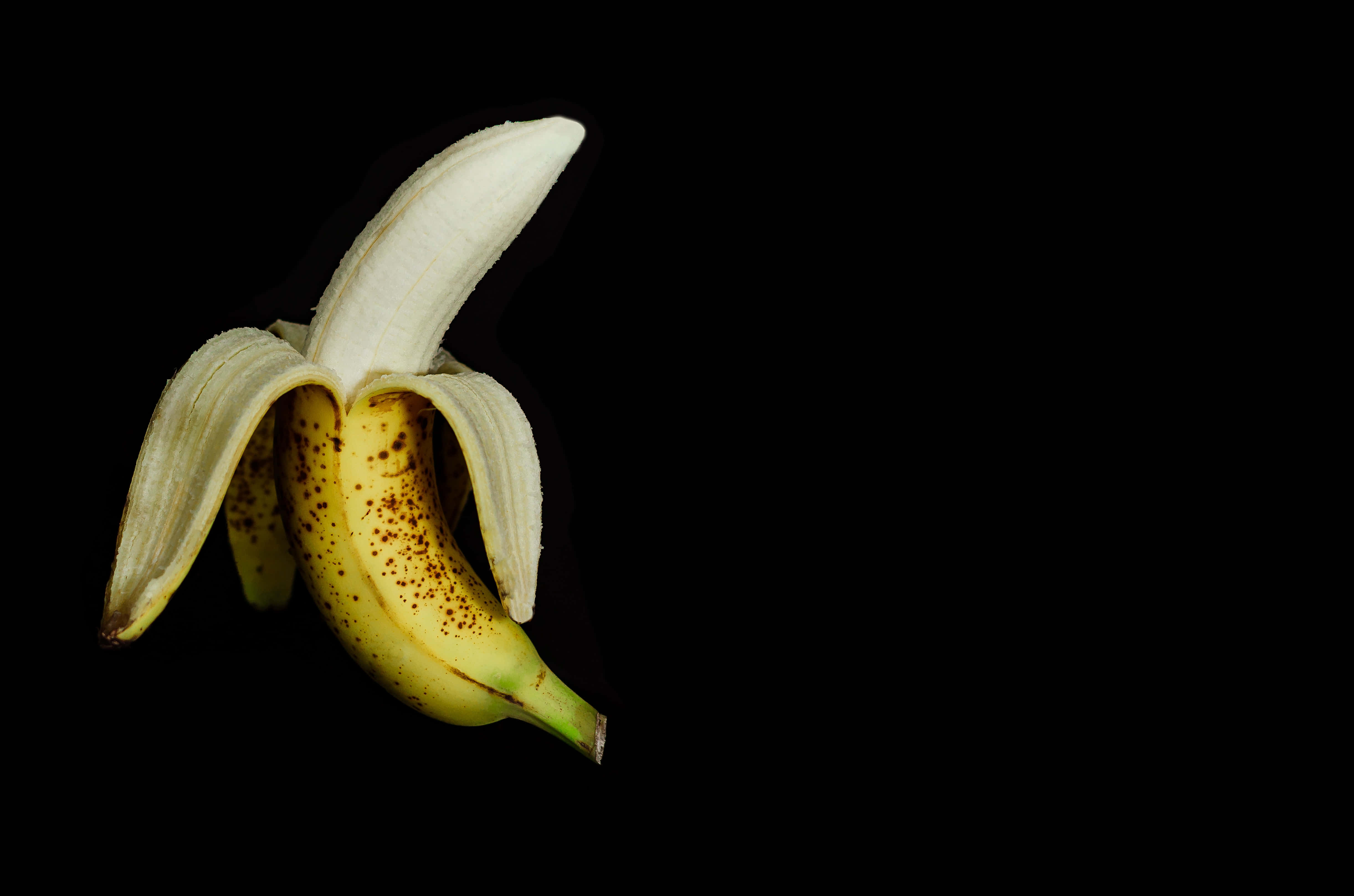 享受多汁美味的香蕉 壁紙 — 一根帶有棕色斑點的成熟香蕉在純黑背景上剝開皮。