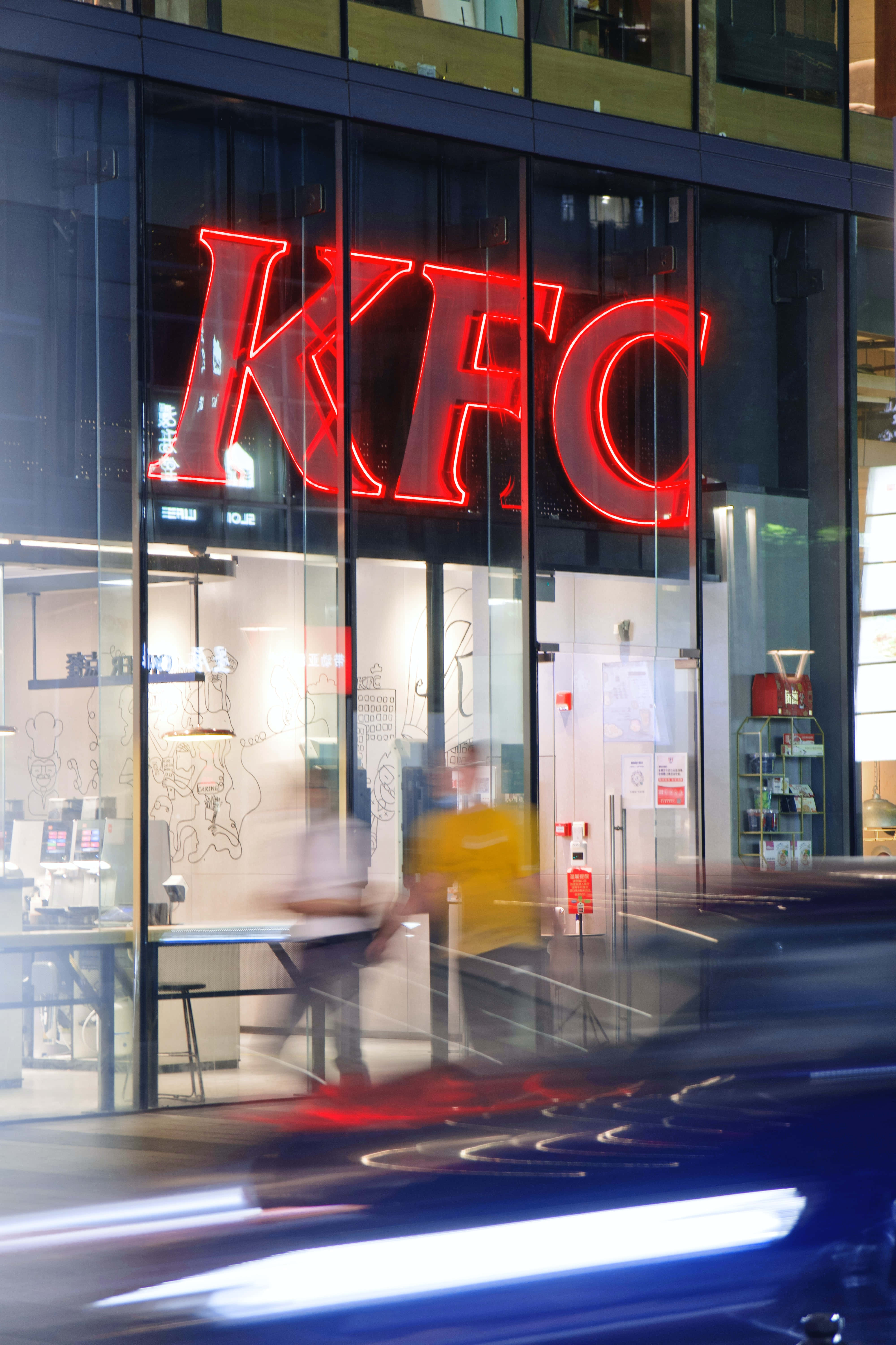 享受您最愛的 KFC 餐點 壁紙 — 一個充滿活力的紅色霓虹燈KFC標誌透過玻璃店面發光，模糊的行人和一輛高速行駛的汽車在夜晚疾馳而過。