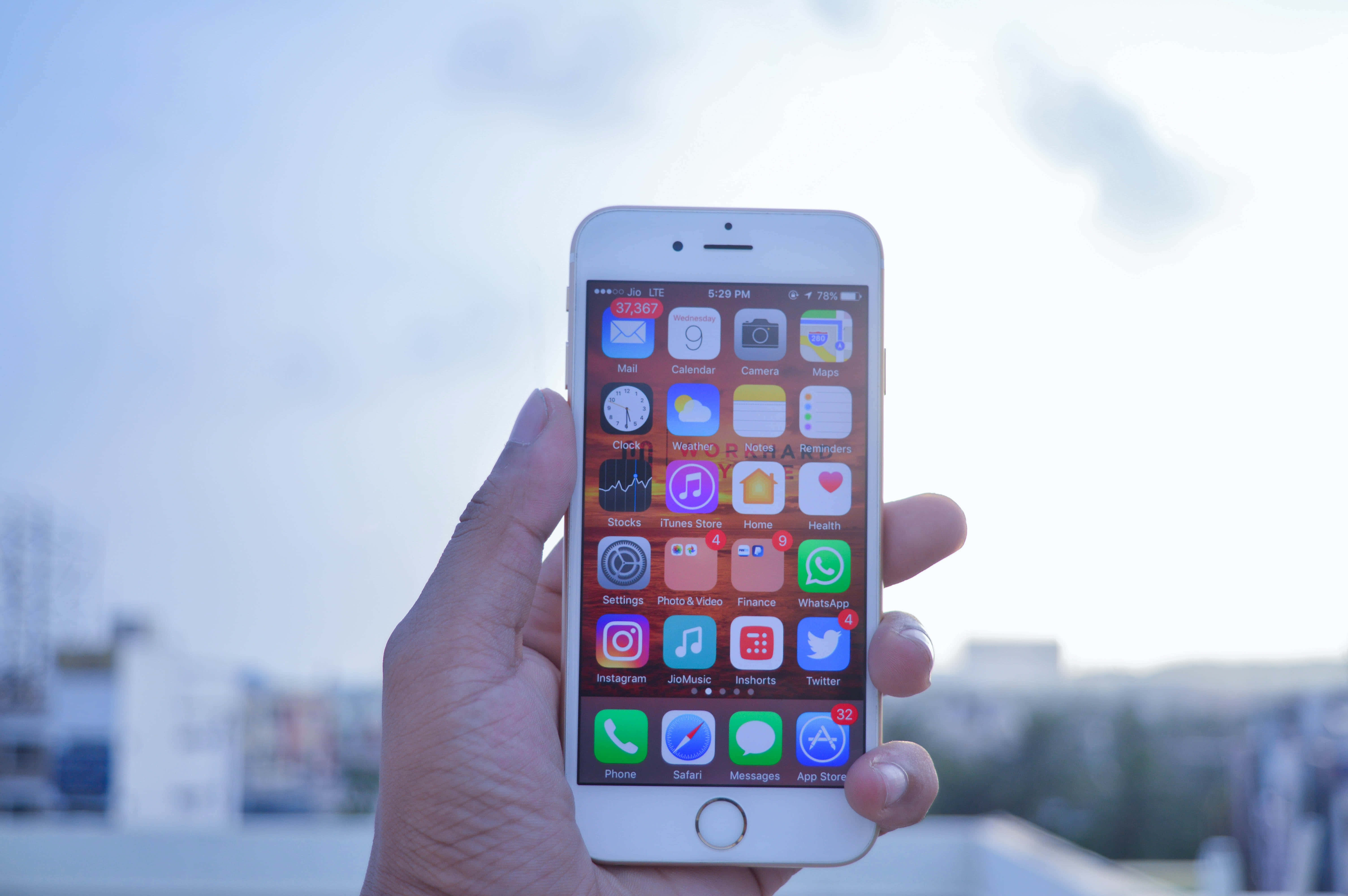 享受最新頂級科技，體驗Apple手機 壁紙 — 一隻手拿著白色iPhone，顯示色彩繽紛的應用程式圖標，背景是明亮朦朧的天空和模糊的城市建築。