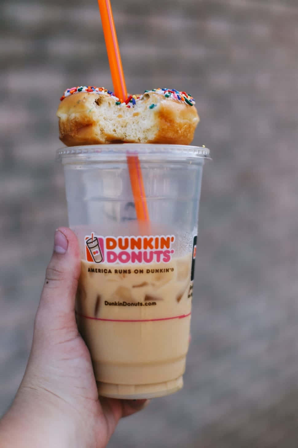 享受清爽冰涼的冰咖啡 壁紙 — 一隻手拿著Dunkin' Donuts冰咖啡，上面放著半個吃剩的彩針甜甜圈和一根橙色吸管。