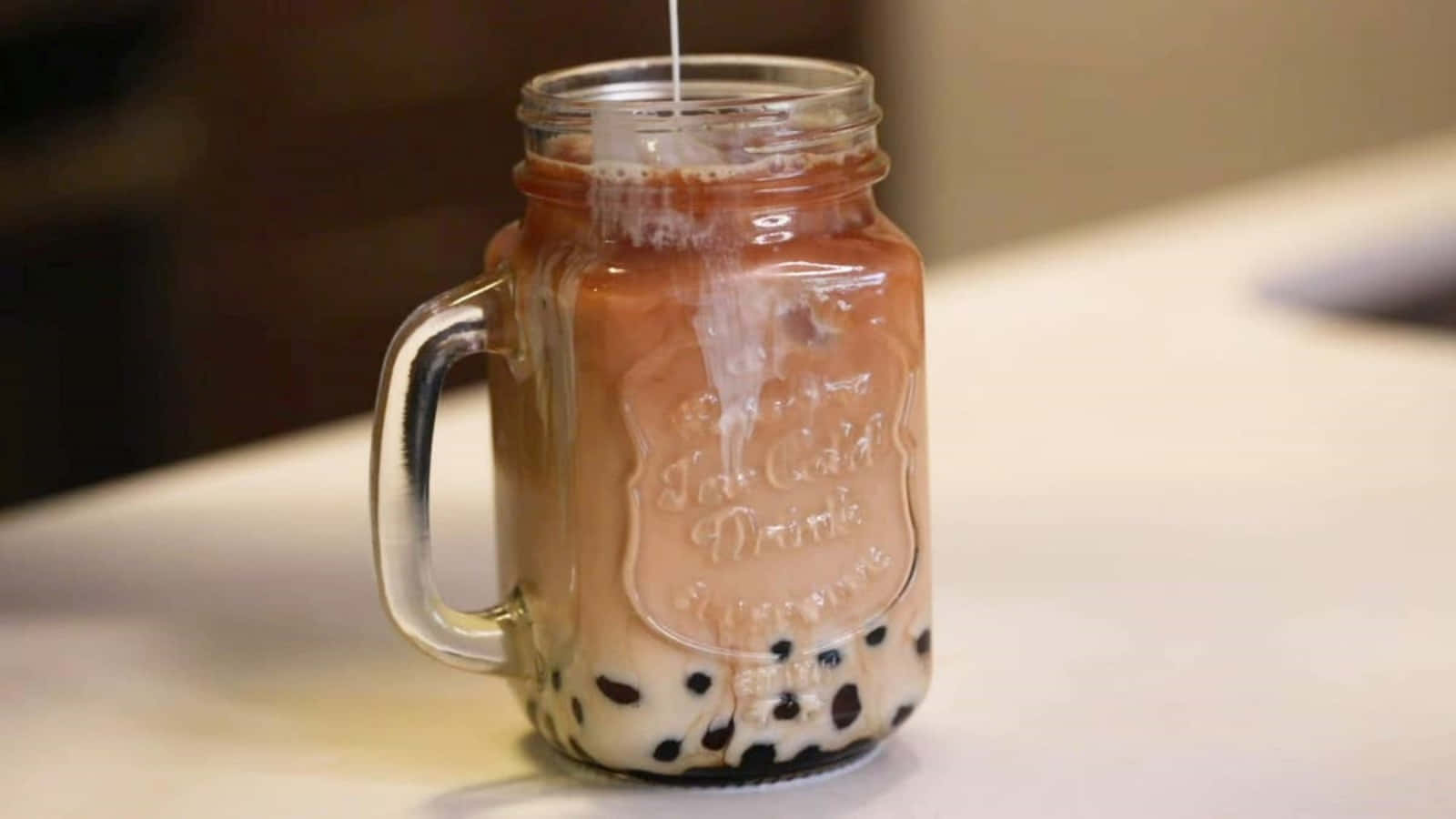 享受清爽珍珠飲品 壁紙 — 一杯帶有黑珍珠的奶油泡泡茶正在倒入白色枱面上的玻璃梅森罐中。