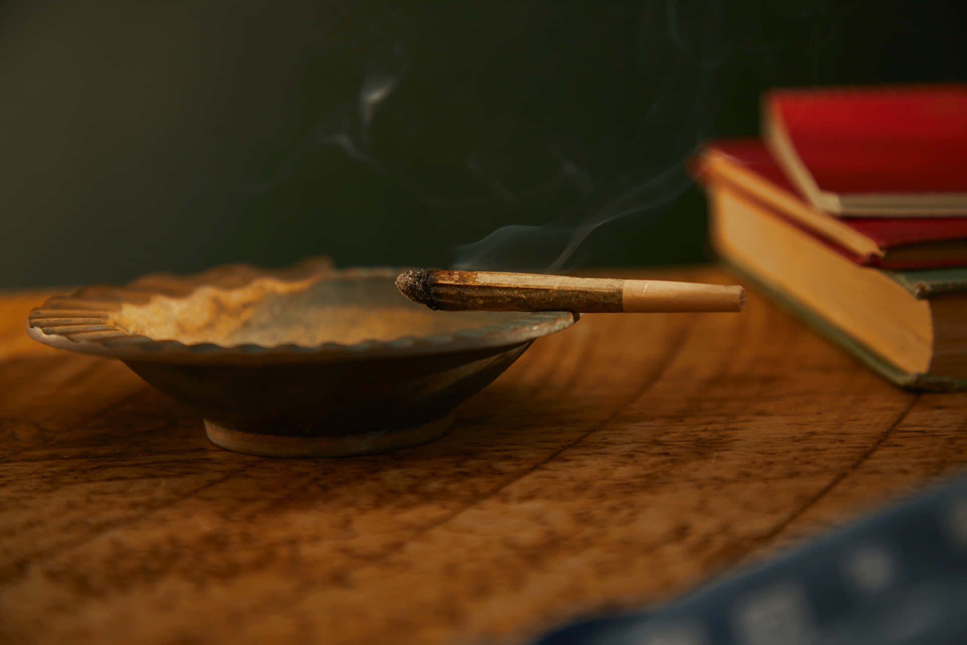 木桌上的煙灰缸裡有一支點燃的雪茄，旁邊有書籍，煙霧上升。