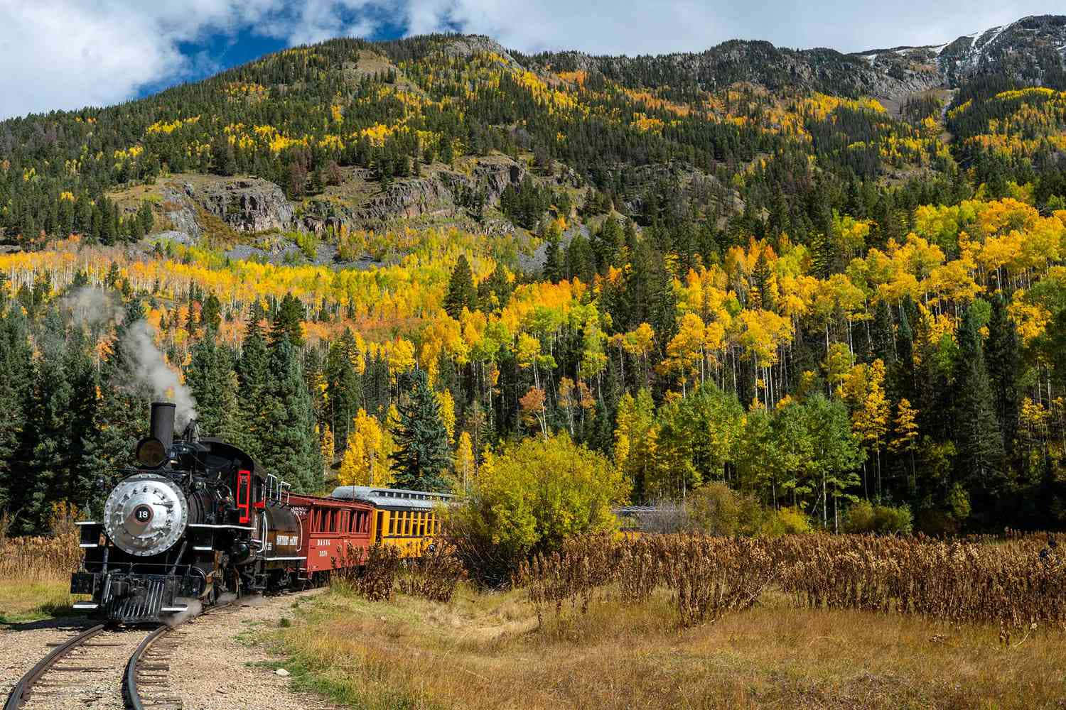 享受科羅拉多巖山的自然美景 壁紙 — 一輛復古蒸汽火車穿過充滿活力的秋日森林，金色白楊和常綠樹在岩石山脈前，天空部分多雲。