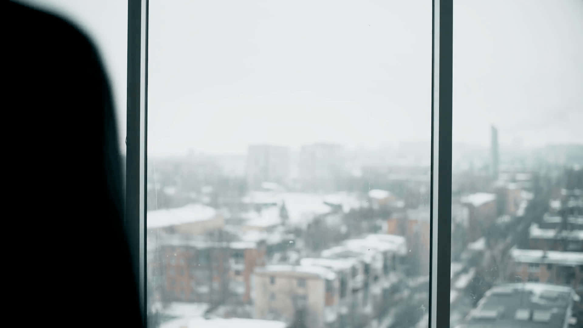 享受窗邊陽光 壁紙 — 一個人的剪影，在陰天時透過窗戶凝視著白雪覆蓋的城市景觀。