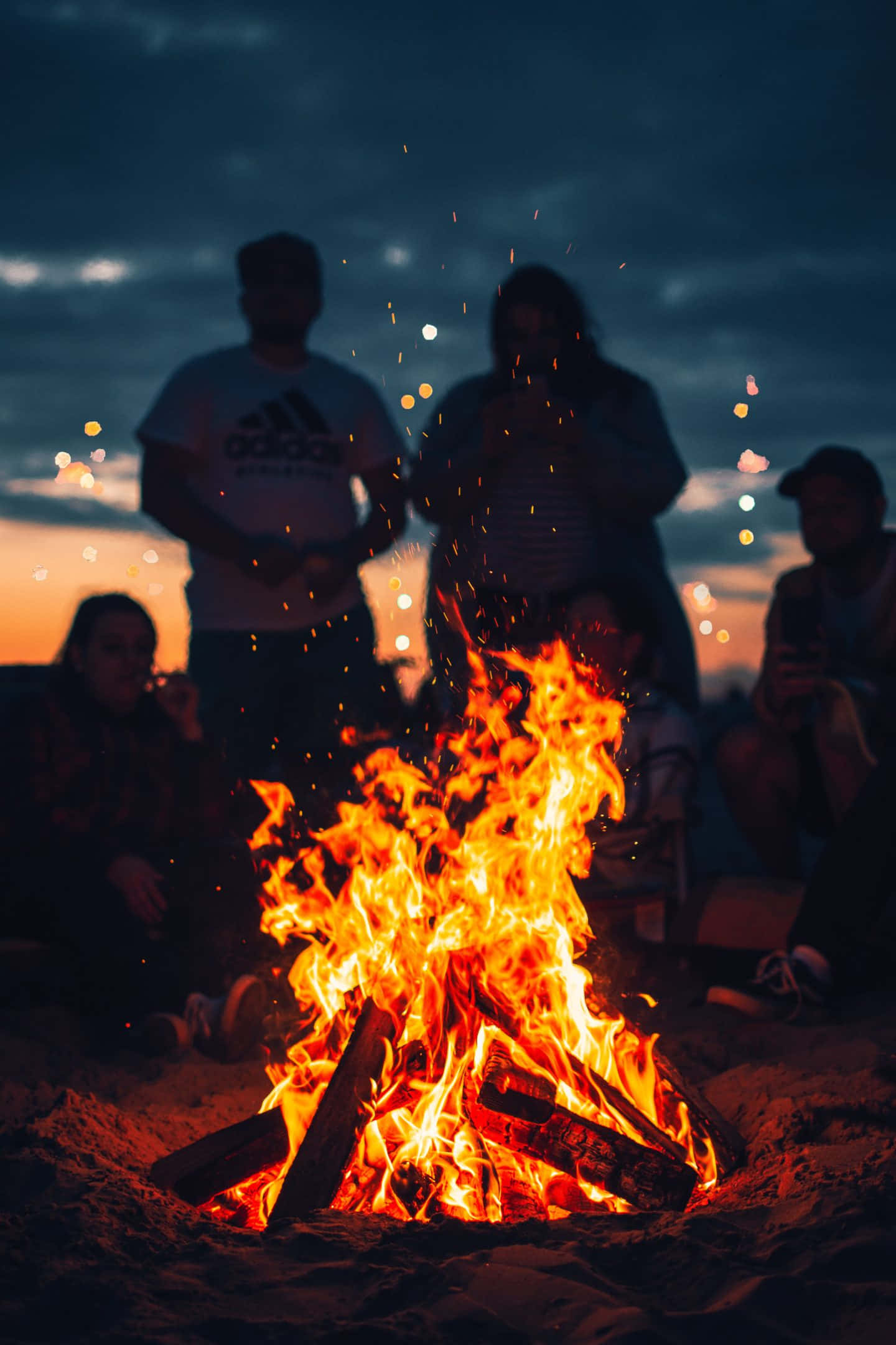 享受篝火旁的夜晚時光 壁紙 — 沙灘上營火噼啪作響，朋友們聚集在暮色天空下，周圍有發光的餘燼和遠處的燈光。