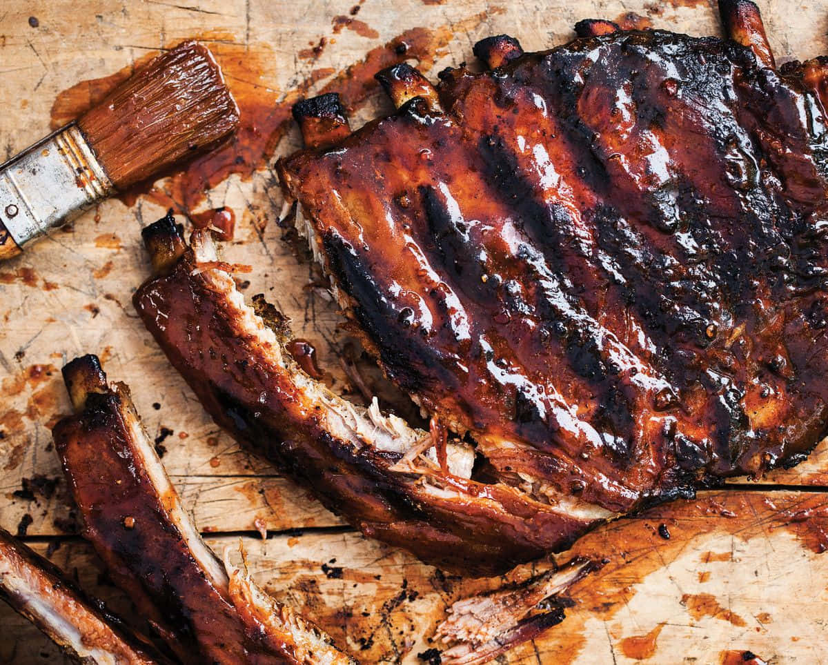 享受美味誘人的BBQ大餐 壁紙 — 閃閃發光的燒烤肋骨帶有焦糖釉面，放在一塊風化的木板上，旁邊有一把用過的塗抹刷。