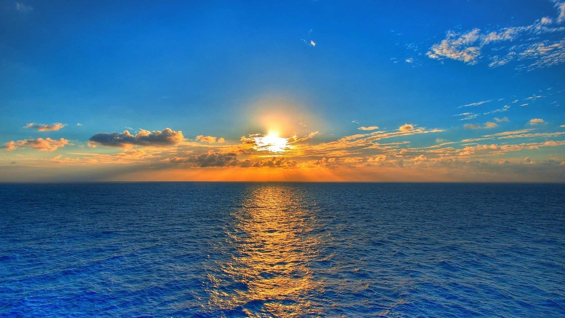 享受美麗陽光 壁紙 — 夕陽映照海洋，金色光芒在波光粼粼的藍色水面上反射，天空色彩斑斕。