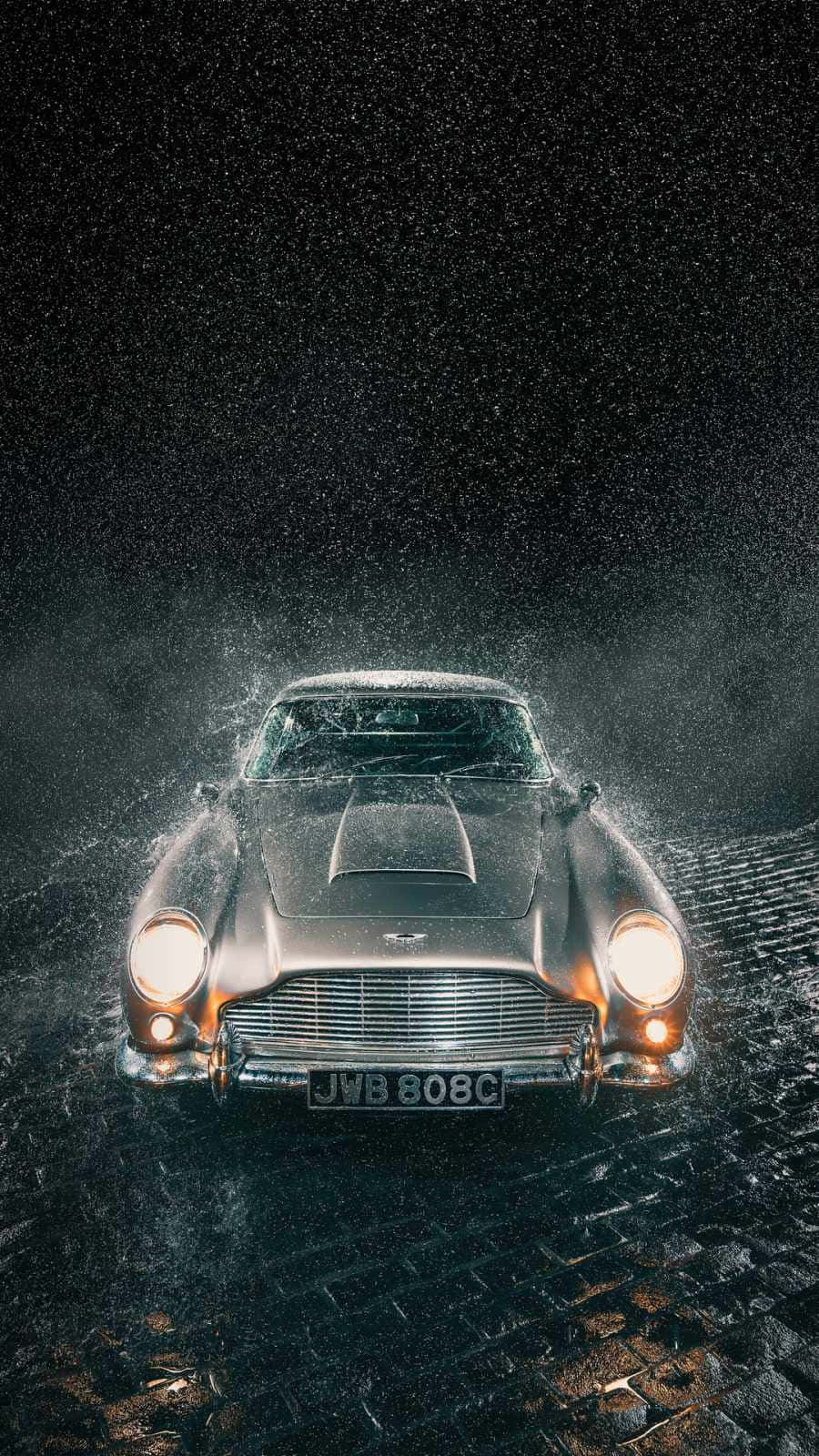 享受阿斯頓·馬丁的奢華駕乘體驗 壁紙 — 一輛銀色Aston Martin DB5從濕滑的鵝卵石中出現，閃爍的車頭燈刺穿黑暗的暴風雨之夜。