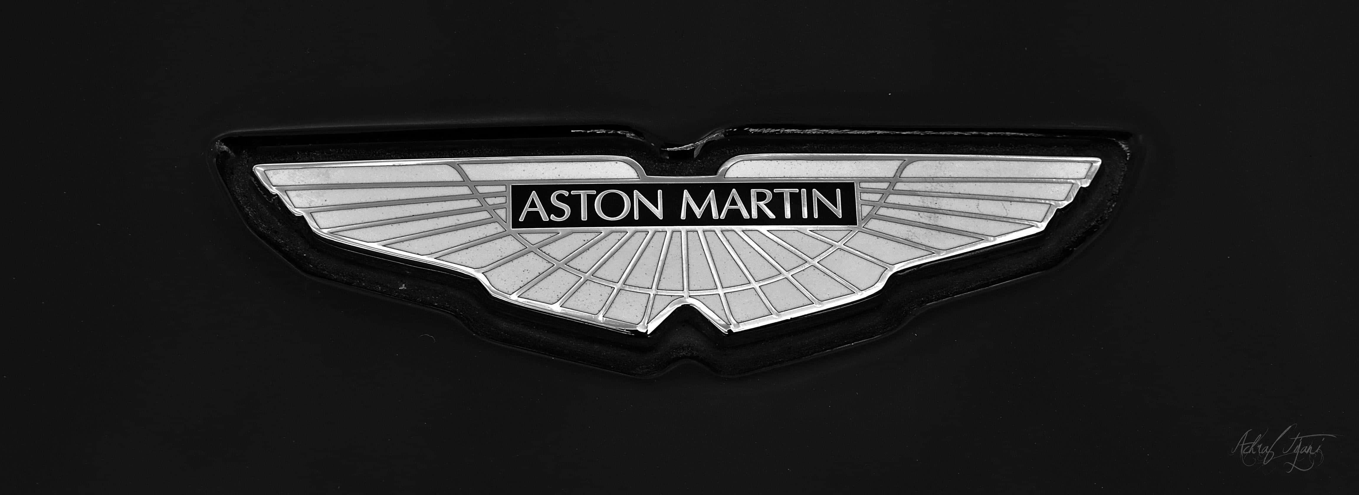 享受阿斯頓·馬丁的奢華體驗 壁紙 — 近距離拍攝銀色Aston Martin翼形標誌徽章在啞光黑色汽車表面上閃耀。