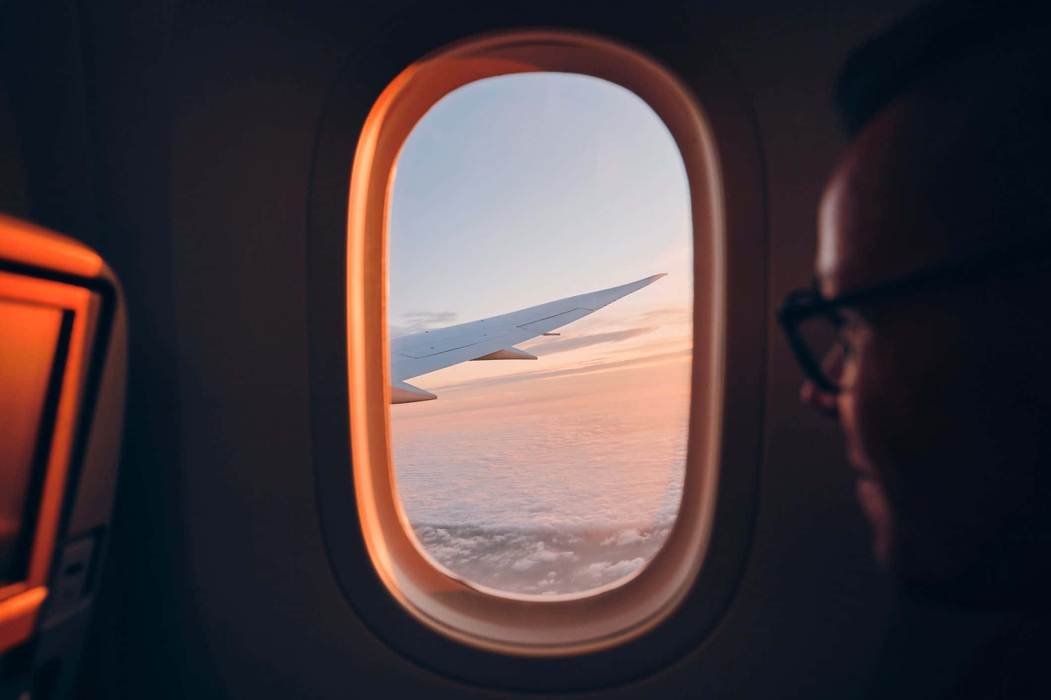 享受飛機窗戶的風景 壁紙 — 一名乘客從飛機窗戶向外凝視，看著機翼劃過沐浴在溫暖日落色調中的雲層。