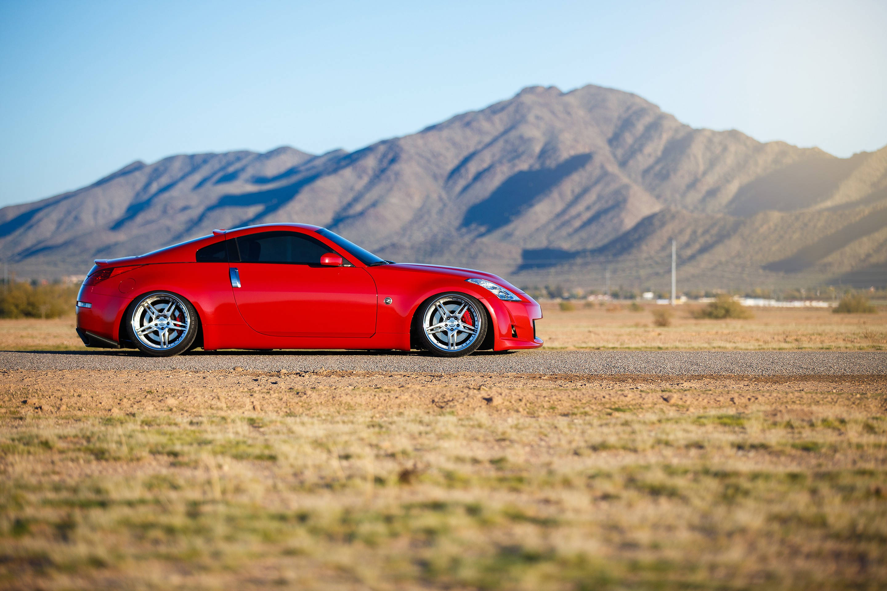 享受駕駛快感，體驗 Nissan 350z 的澎湃動力 壁紙 — 一輛鮮紅色跑車停泊在沙漠公路上，背景是崎嶇的山脈，天空清澈湛藍。
