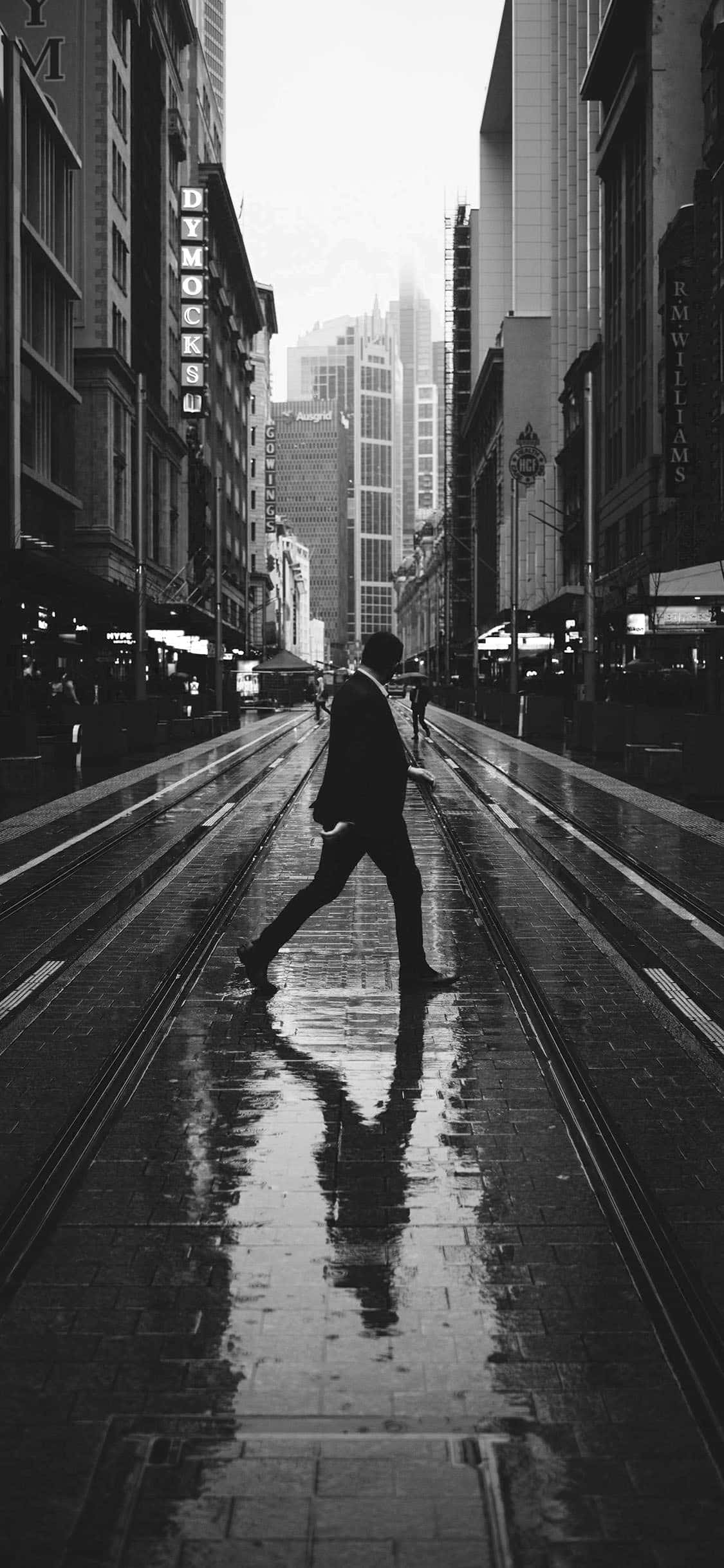 享受黑白 iPhone 的經典優雅 壁紙 — 一個孤獨的身影自信地走在被雨水浸濕的城市街道上，兩旁是高聳的建築物，霓虹燈招牌以單色發光。