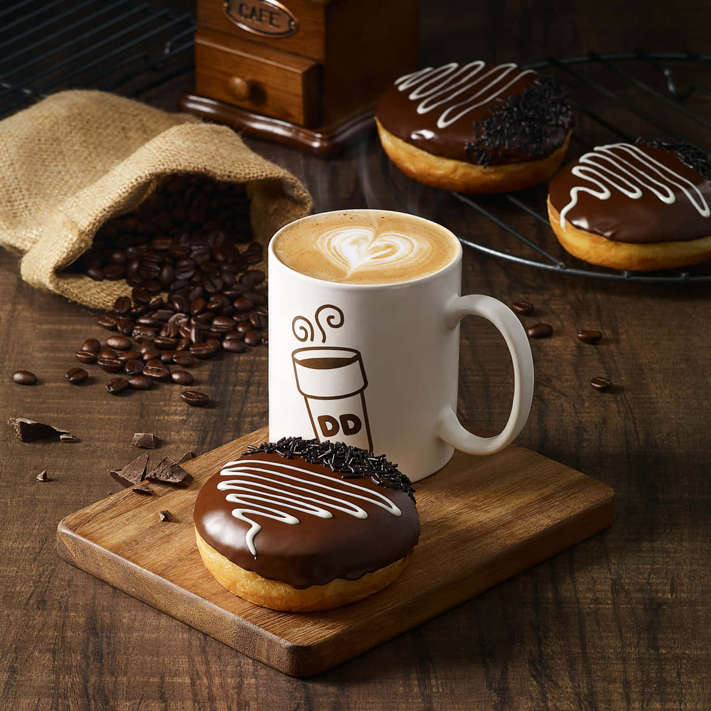 享用 Dunkin' Donuts 的美味咖啡 壁紙 — 一杯帶有心形拉花的熱拿鐵，旁邊是沾有朱古力的冬甩，放在一塊被咖啡豆和麻布袋圍繞的木板上。