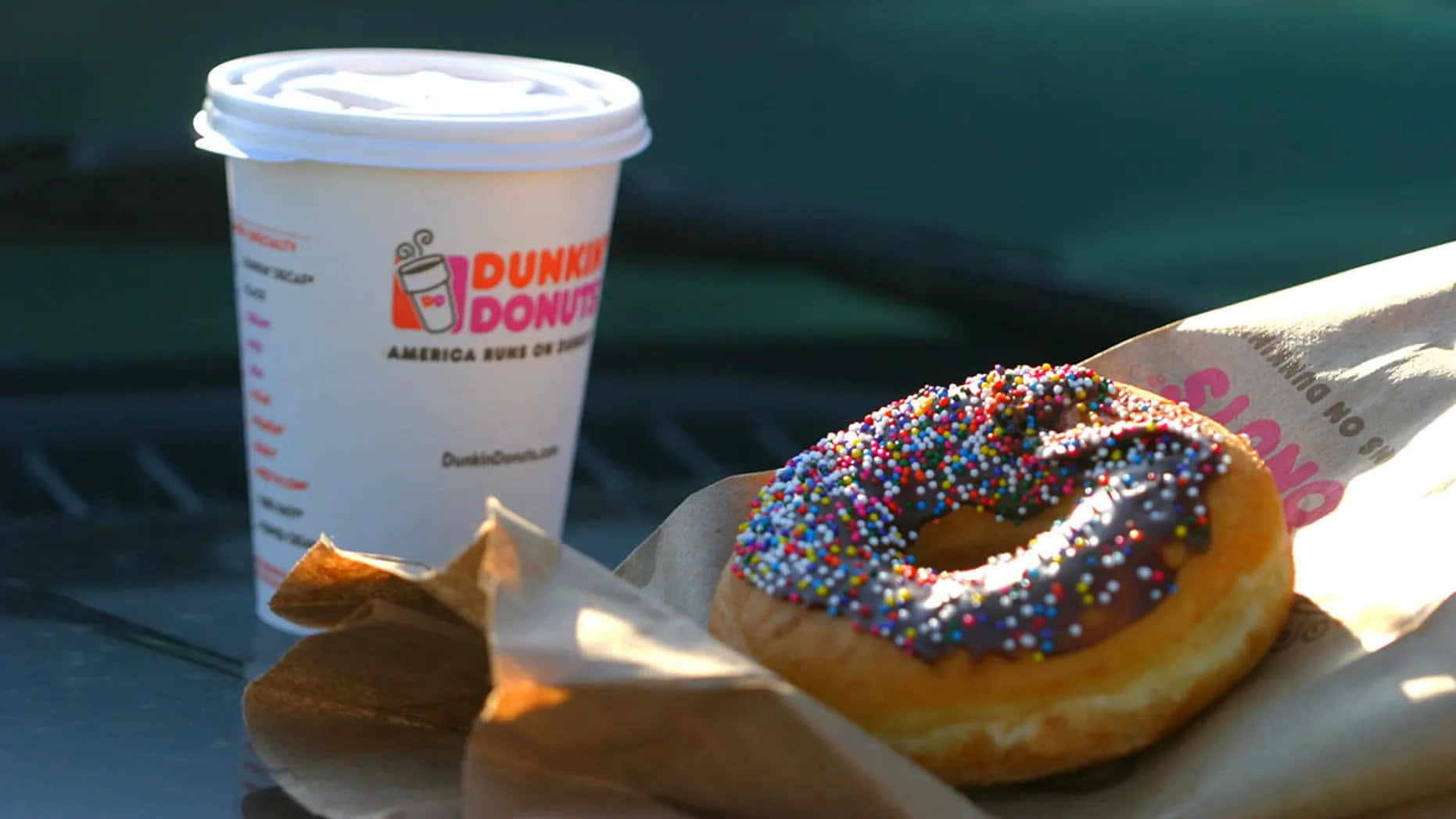 享用Dunkin'新鮮製作的美味甜甜圈 壁紙 — 一杯Dunkin' Donuts咖啡和一個朱古力碎甜甜圈放在溫暖陽光照射的深色表面上。