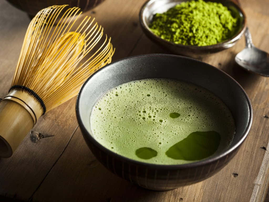 享用一杯抹茶 壁紙 — 一碗充滿泡沫的鮮綠色抹茶旁邊放著竹筅和粉末，置於質樸的木桌上。