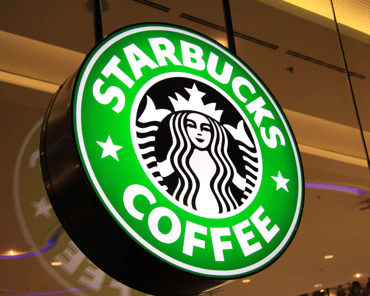 享用一杯星巴克咖啡，暫時放鬆一下 壁紙 — 一個明亮照明的Starbucks咖啡標誌牌掛在室內，發出綠色光芒，帶有白色文字和人魚標誌。