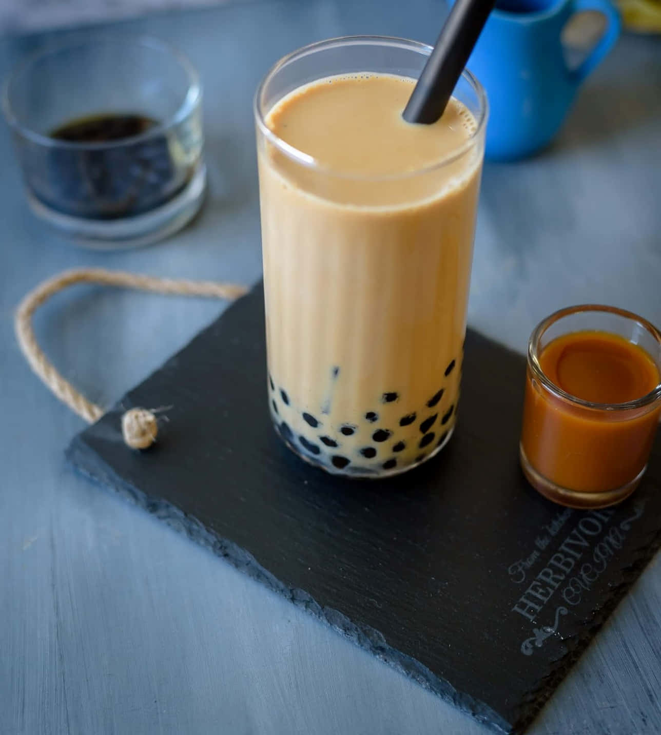 享用一杯美味的珍珠奶茶 壁紙 — 一杯高身嘅忌廉泡泡茶配黑色木薯珍珠，放喺石板托盤上，旁邊有焦糖醬同藍色杯。