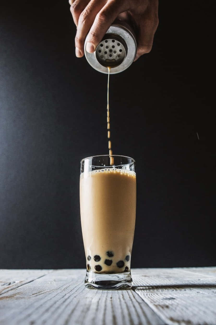 享用果味波霸奶茶，香甜可口 壁紙 — 一隻手將金屬搖杯中的奶油泡沫奶茶倒入裝有木薯珍珠的玻璃杯中，放在質樸的木質表面上。