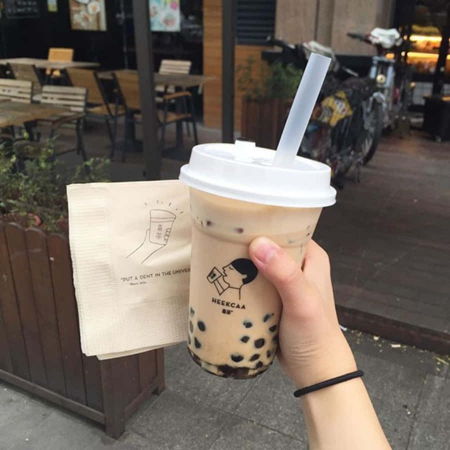 享用清爽珍珠奶茶 壁紙 — 一隻手喺戶外咖啡廳座位區附近，拎住一杯奶油味珍珠奶茶（有黑色木薯珍珠）同一張有品牌嘅紙巾。