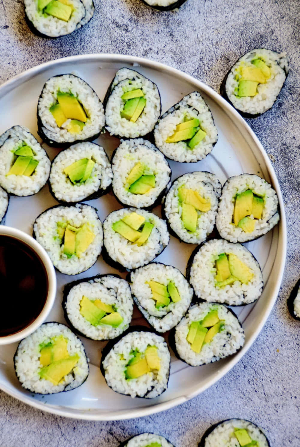 享用美味壽司捲 壁紙 — 新鮮牛油果壽司卷放在白色碟上，旁邊有醬油，置於有紋理的灰色表面上。