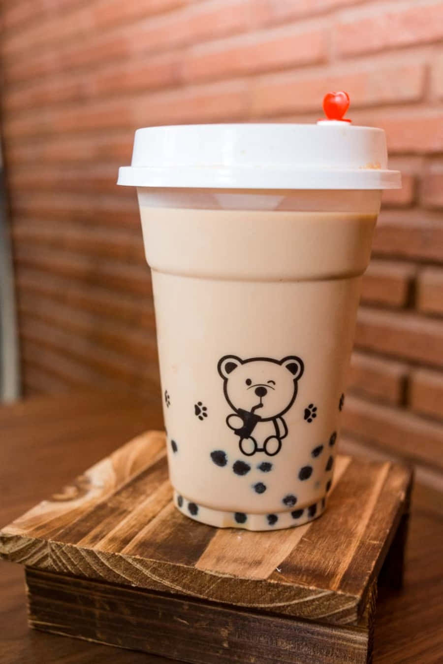 享用美味珍珠奶茶 壁紙 — 一杯帶有熊仔設計的白色杯裝忌廉泡泡茶，放在木架上，背景係溫暖嘅磚牆。