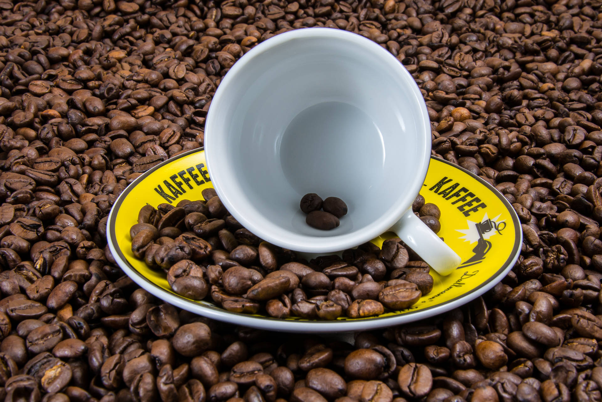 白色咖啡杯和碟子，黃色邊緣上有 'KAFFEE' 文字，周圍環繞著烤咖啡豆。