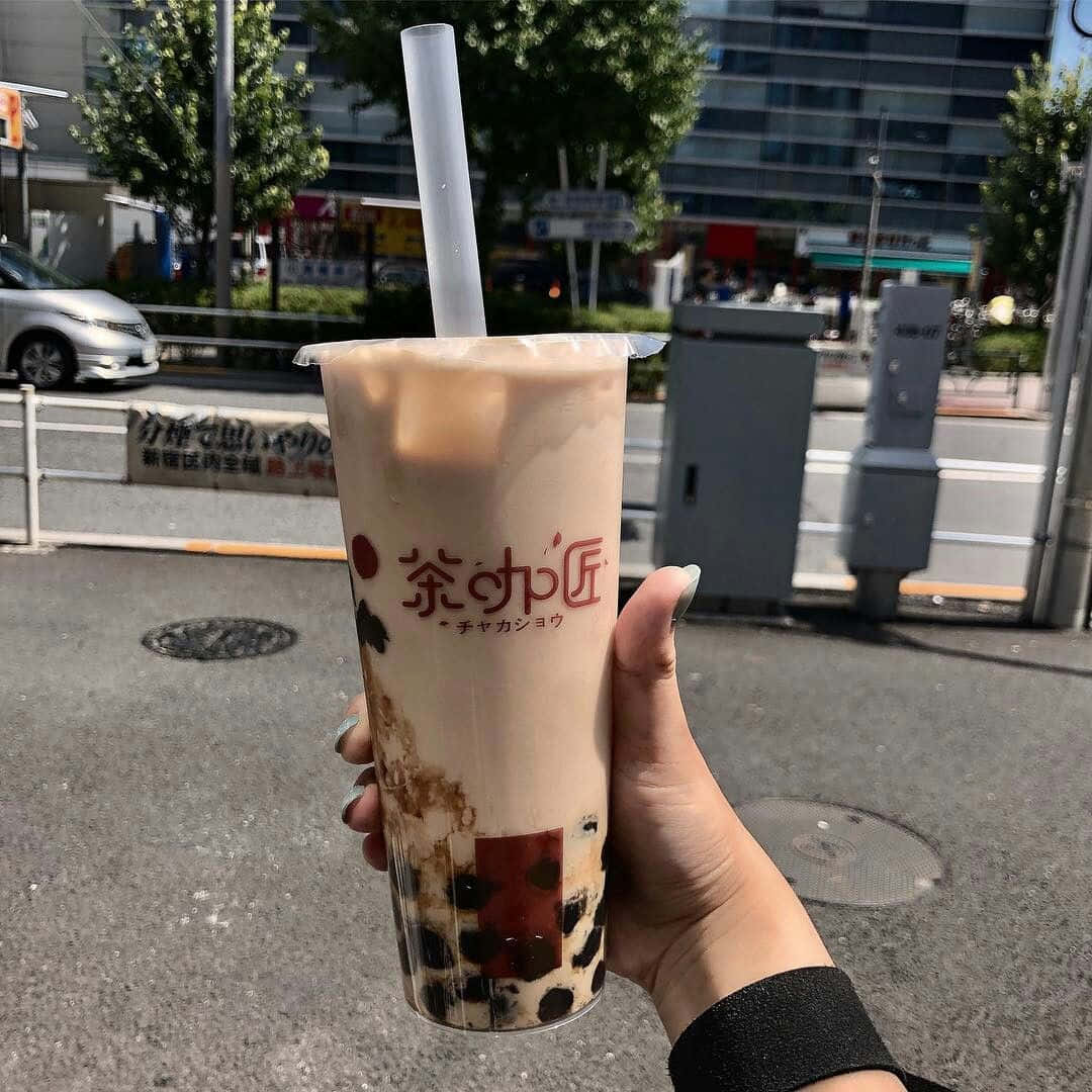 享用香甜珍珠奶茶 壁紙 — 一隻手喺明亮嘅城市街道背景下，揸住一杯有黑色珍珠嘅忌廉泡泡茶。