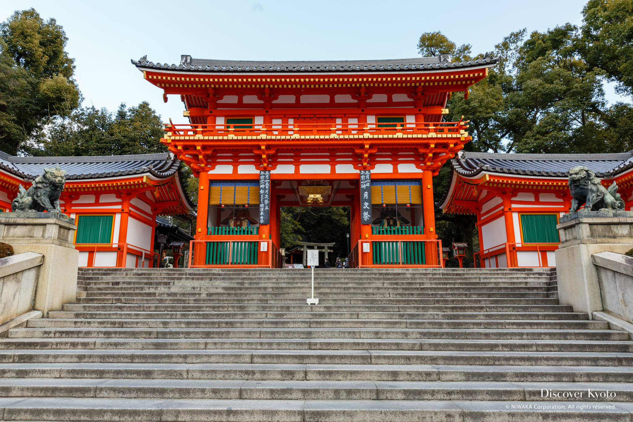 京都八坂神社 壁紙 — 一座宏偉的傳統日本神社，擁有鮮豔的橙色鳥居和石階，兩側有守護獅子雕像，天空晴朗。