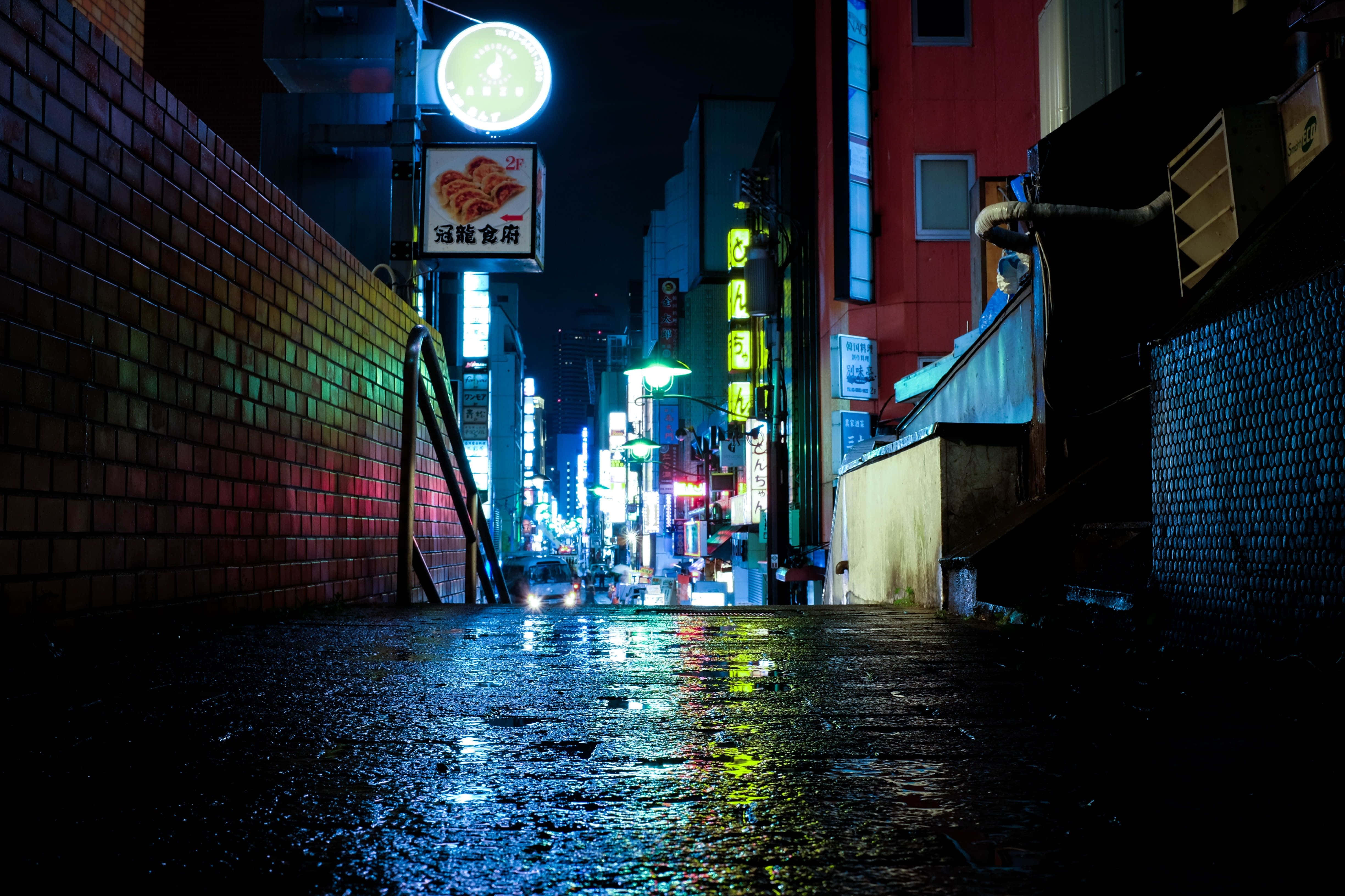 京都日本夜景，紅色霓虹招牌主宰的璀璨夜色 壁紙 — 雨濕的小巷在夜晚反射高聳建築物的霓虹招牌，營造出繁華的市區景象。