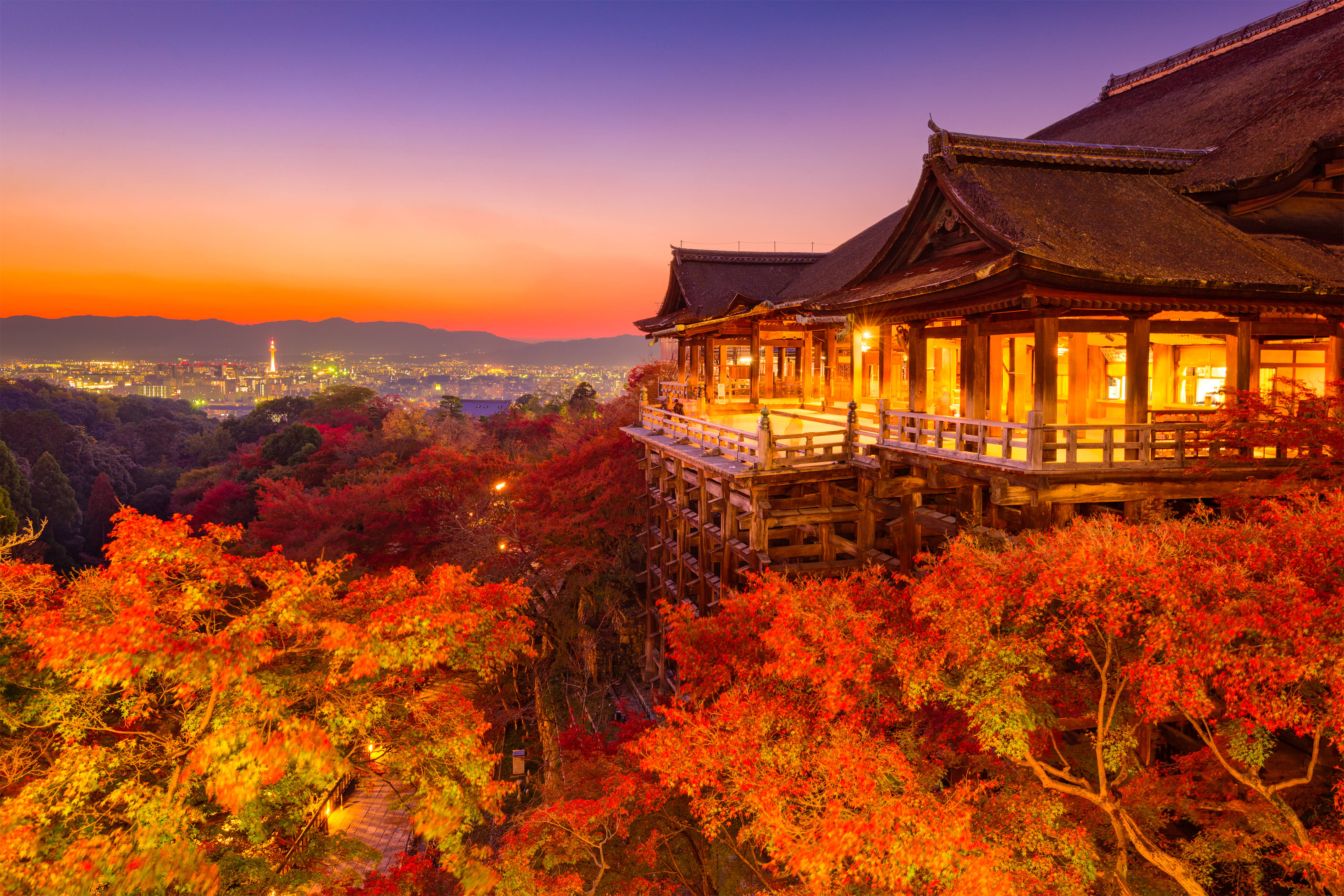京都清水寺秋景 壁紙 — 一座傳統木造寺廟坐落在山坡上，周圍環繞著火紅的秋葉，暮色中下方是閃爍的城市景觀。