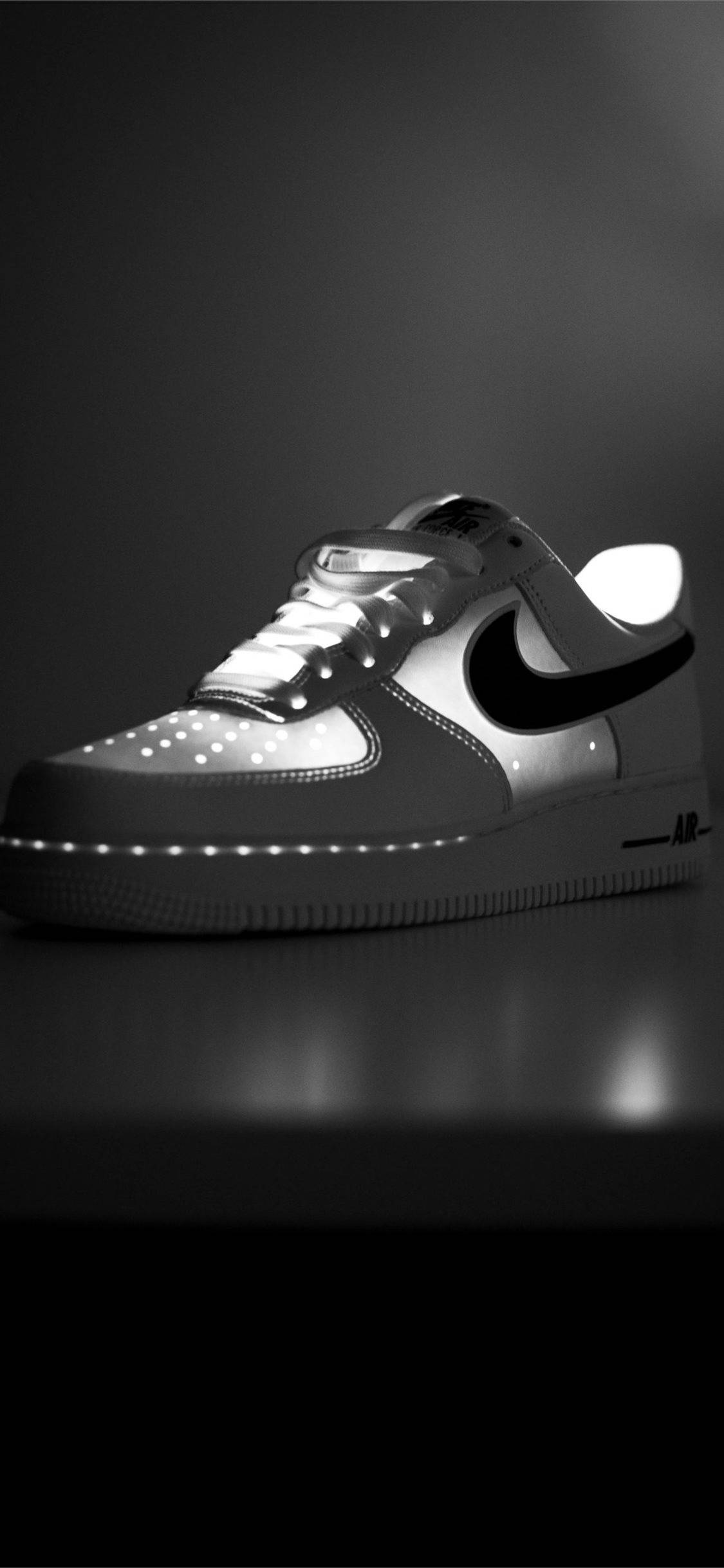 亮燈 Nike iPhone 簡約款 壁紙 — 一雙單色Nike Air Force 1運動鞋，鞋帶和鞋底帶有發光LED燈，放置在反光表面上，背景為深色漸變。