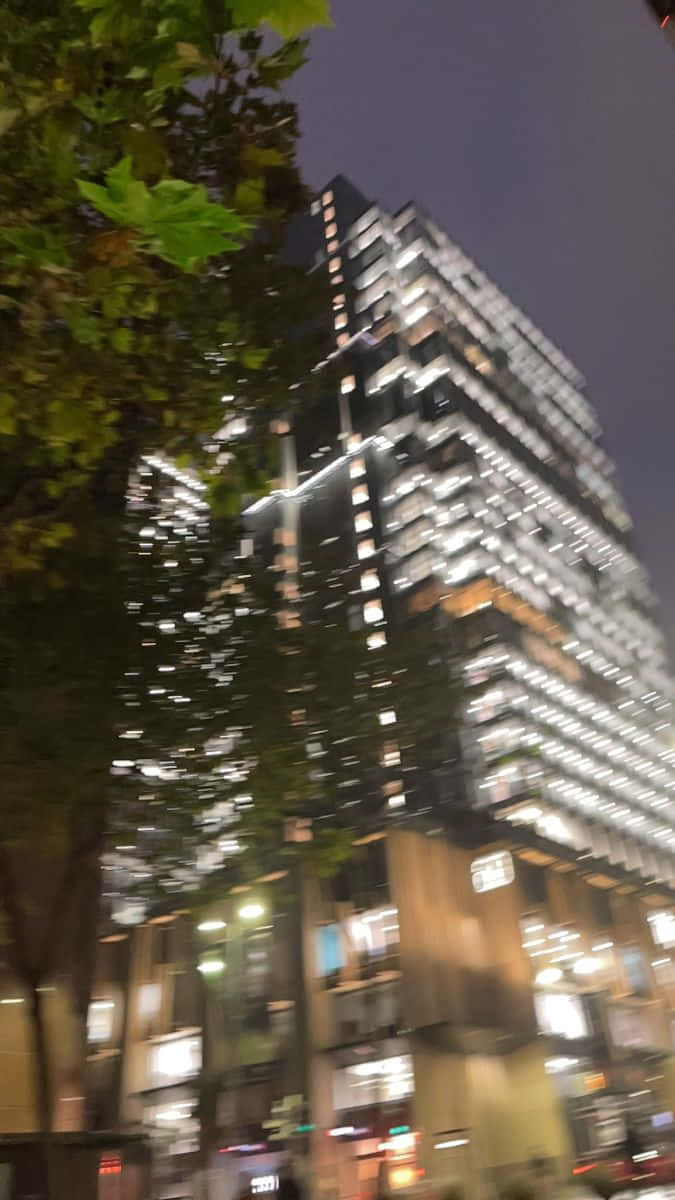 夜晚亮燈的高聳玻璃建築，前景有模糊的樹木。