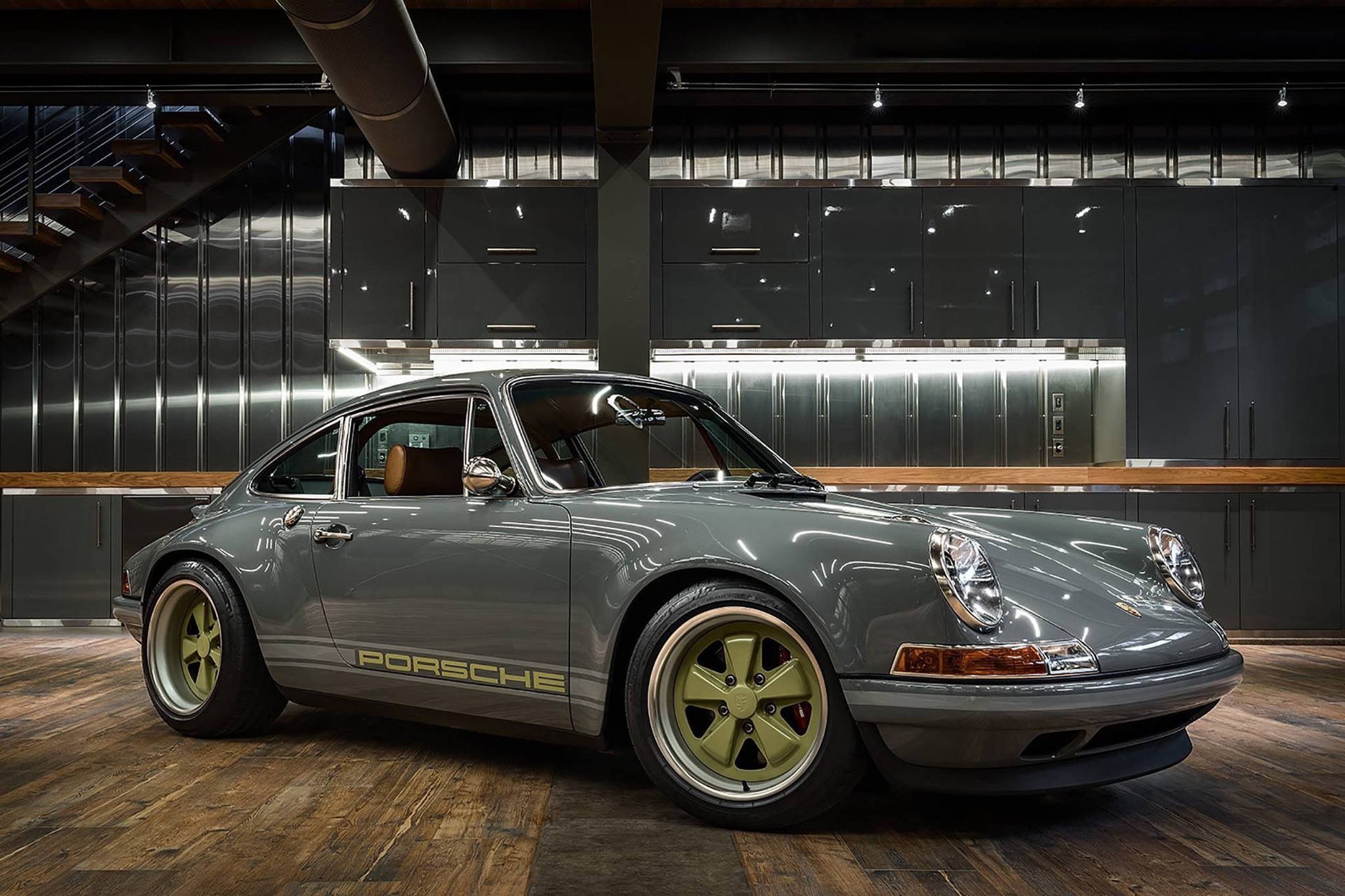 亮面灰Singer Porsche 壁紙 — 一輛時尚的灰色保時捷911配金色輪圈，在現代工業車庫的攝影棚燈光下閃閃發光，車庫內有深色櫥櫃和木地板。