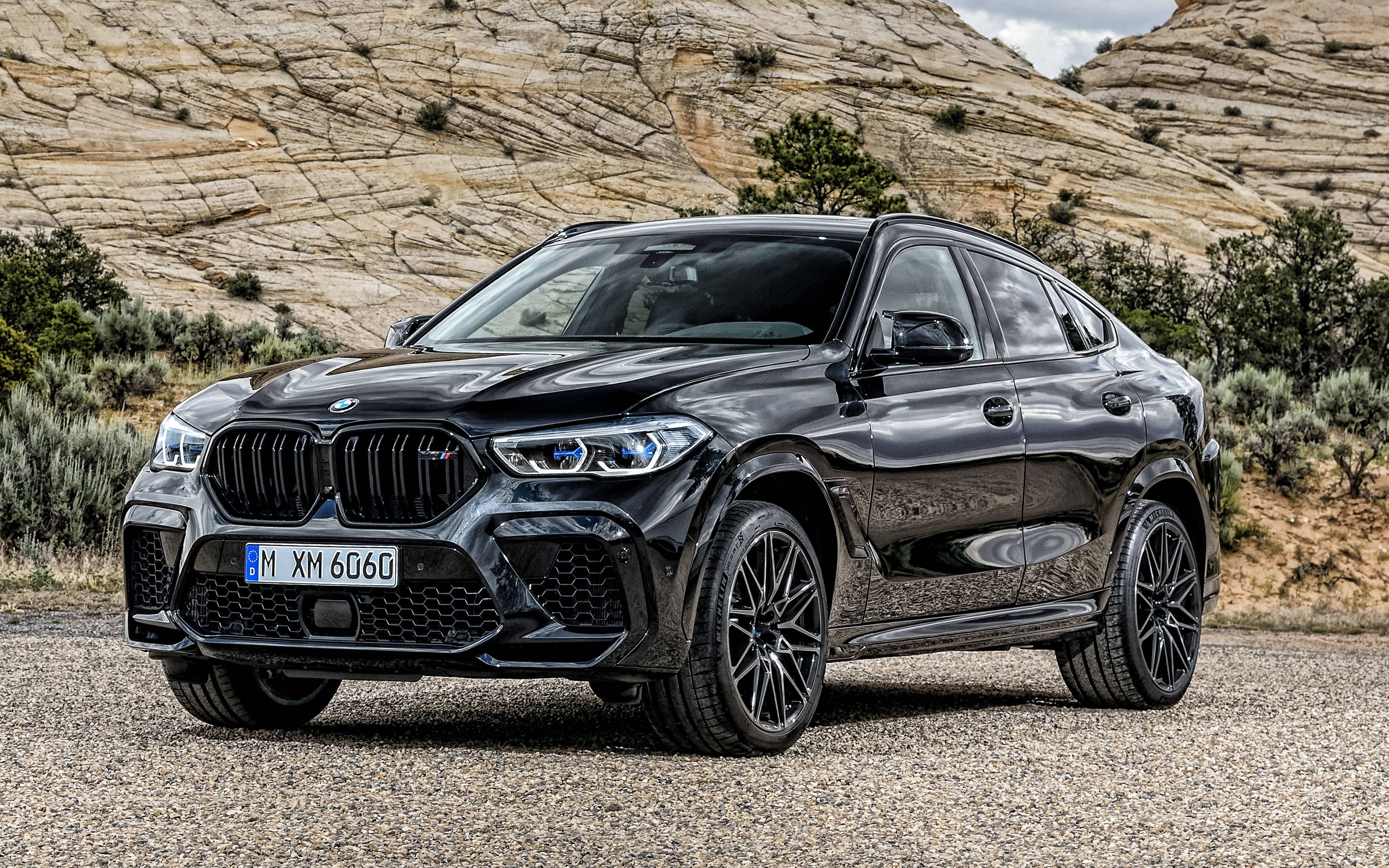亮面黑色 BMW X6 M 壁紙 — 一輛亮黑色BMW X6 M停在礫石上，背景有堅固的岩層和稀疏的沙漠植被。
