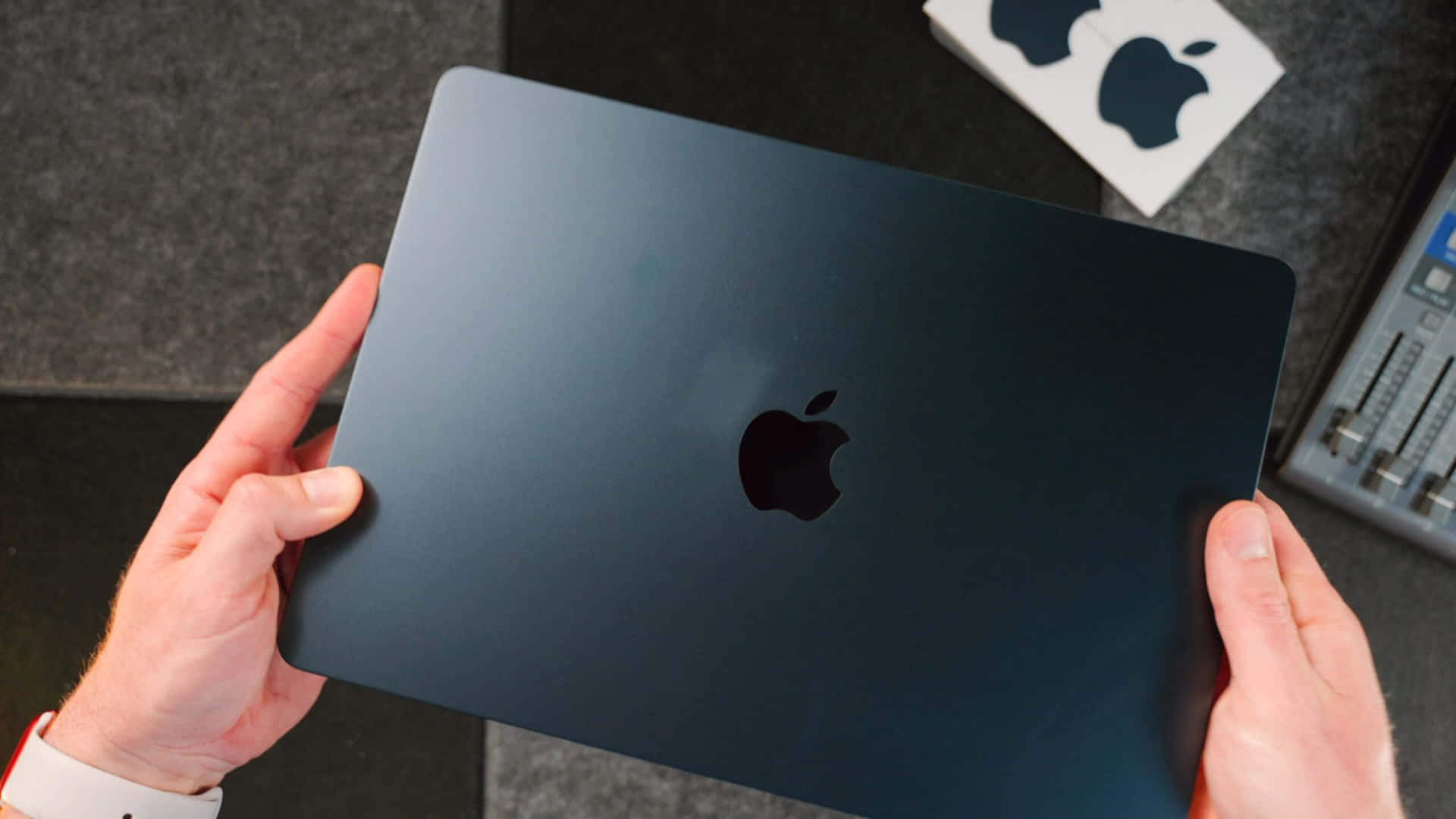 一個人在深色桌面上拿著黑色 MacBook，旁邊有貼紙和音響設備。