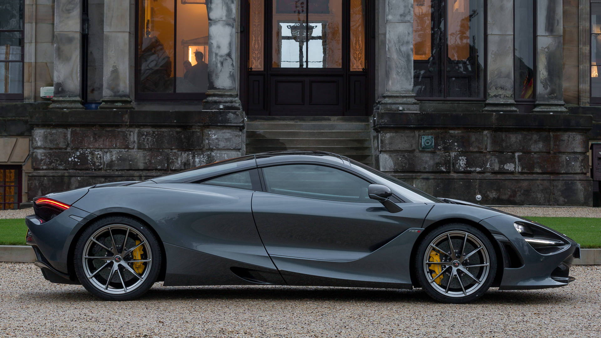 亮面黑色麥拉倫720S 4K 壁紙 — 一架流線型啞光灰色McLaren 720S跑車停泊在碎石上，前面係一座有溫暖室內燈光嘅大型石建築。